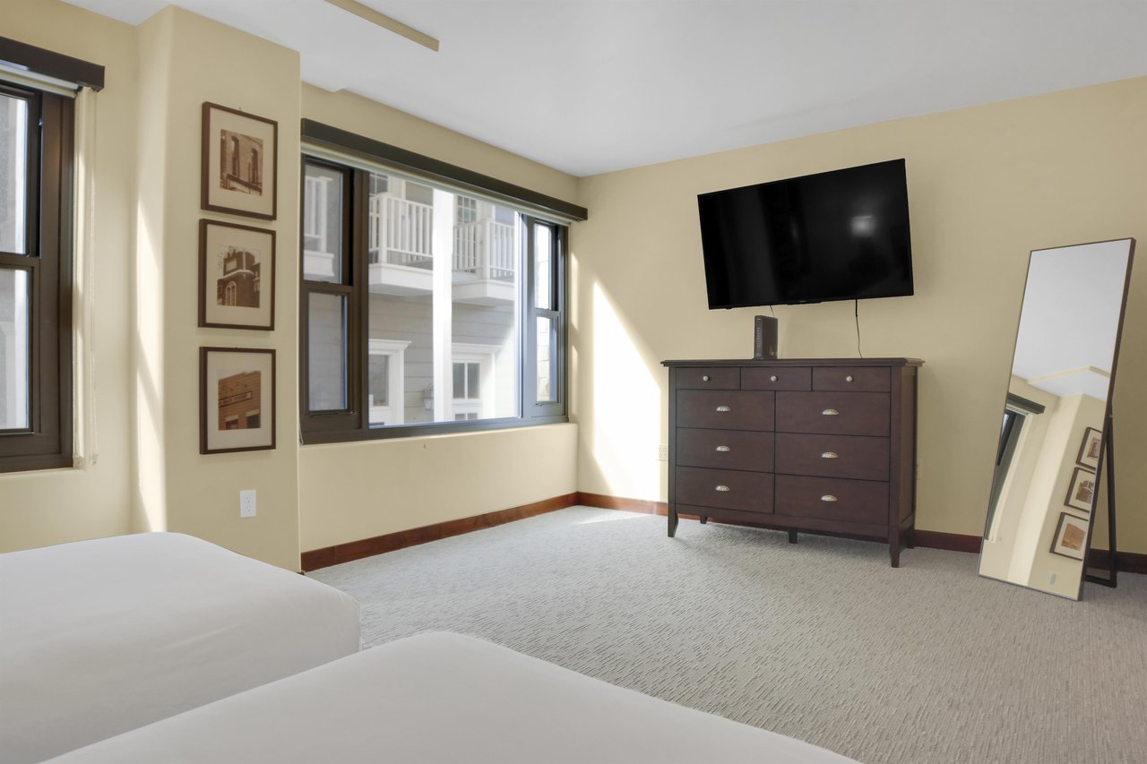The end of two twin beds with a wooden dresser and wall mounted TV, large picture window and cream colored carpet.