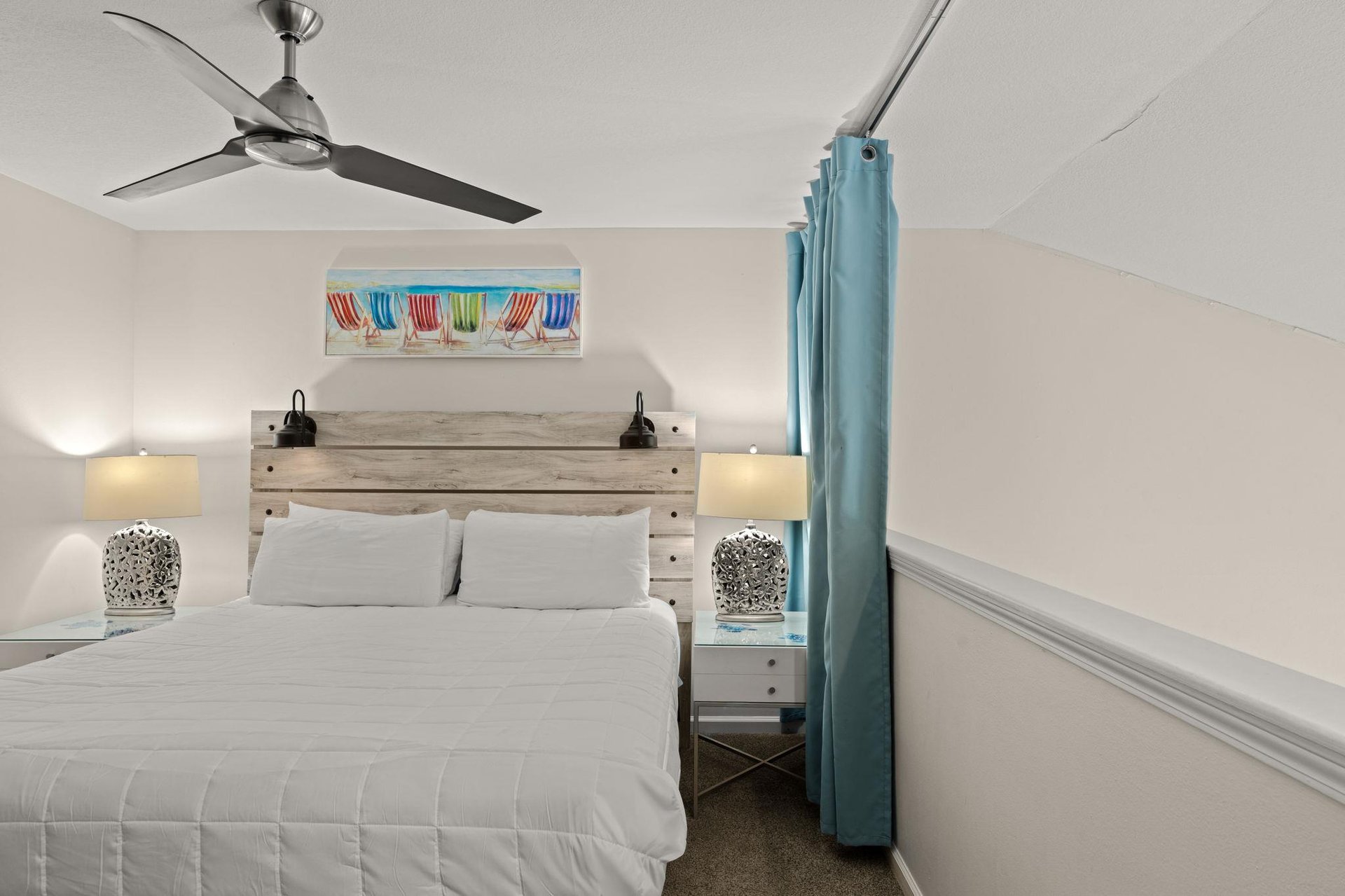 Unwind in your cozy coastal retreat with a plush bed, whimsical beach chair art, and soothing bedside lamps. The ceiling fan keeps you comfortable as ocean breezes drift through.