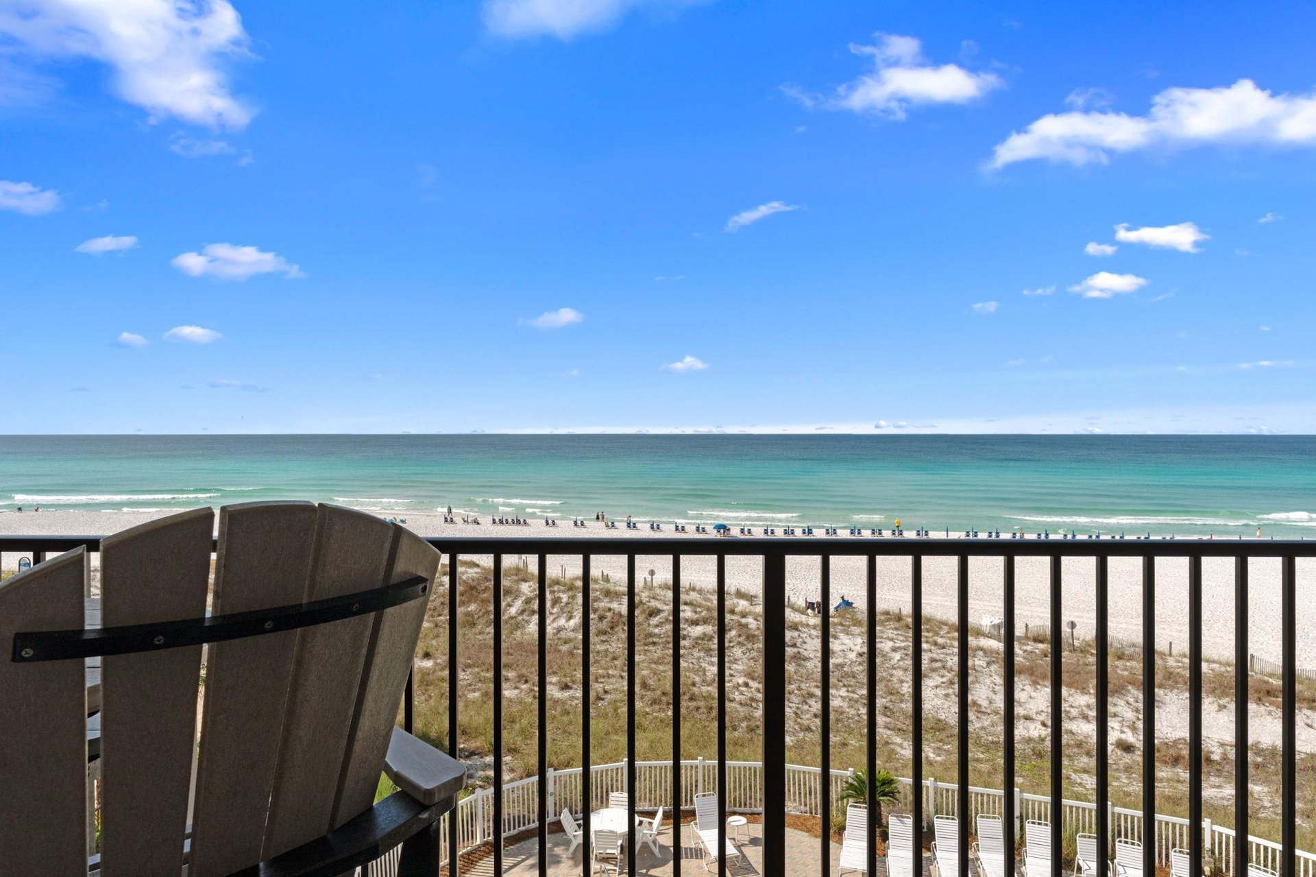 Beachfront balcony overlooks white sand shores and emerald waters under bright blue skies, with comfortable seating to watch dolphins play and sunsets paint the horizon.