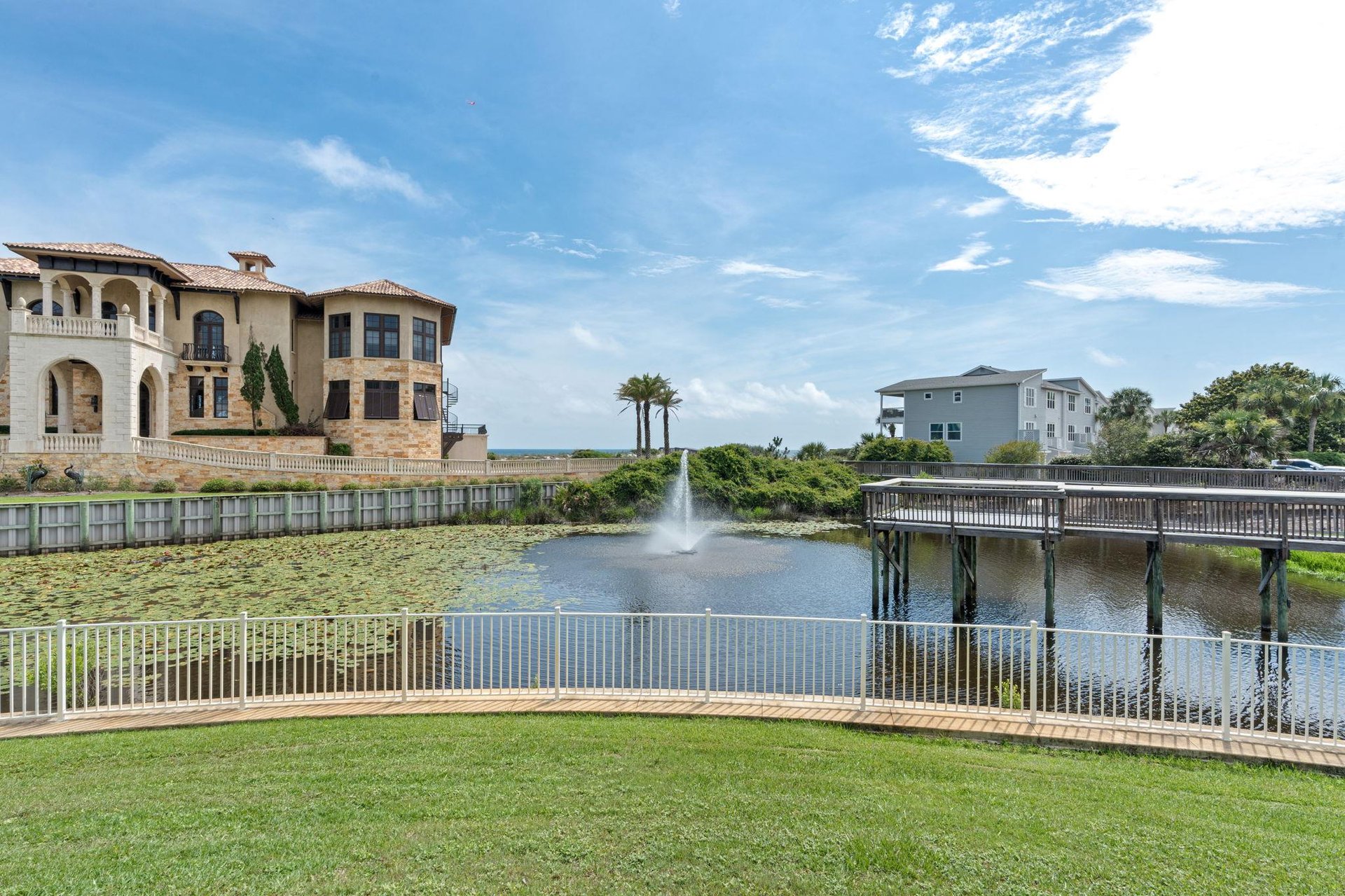 Vacation rental property surrounded by tranquil waterfront community with landscaped pond, fountain, walking bridge, and nearby neighboring homes under clear blue skies.