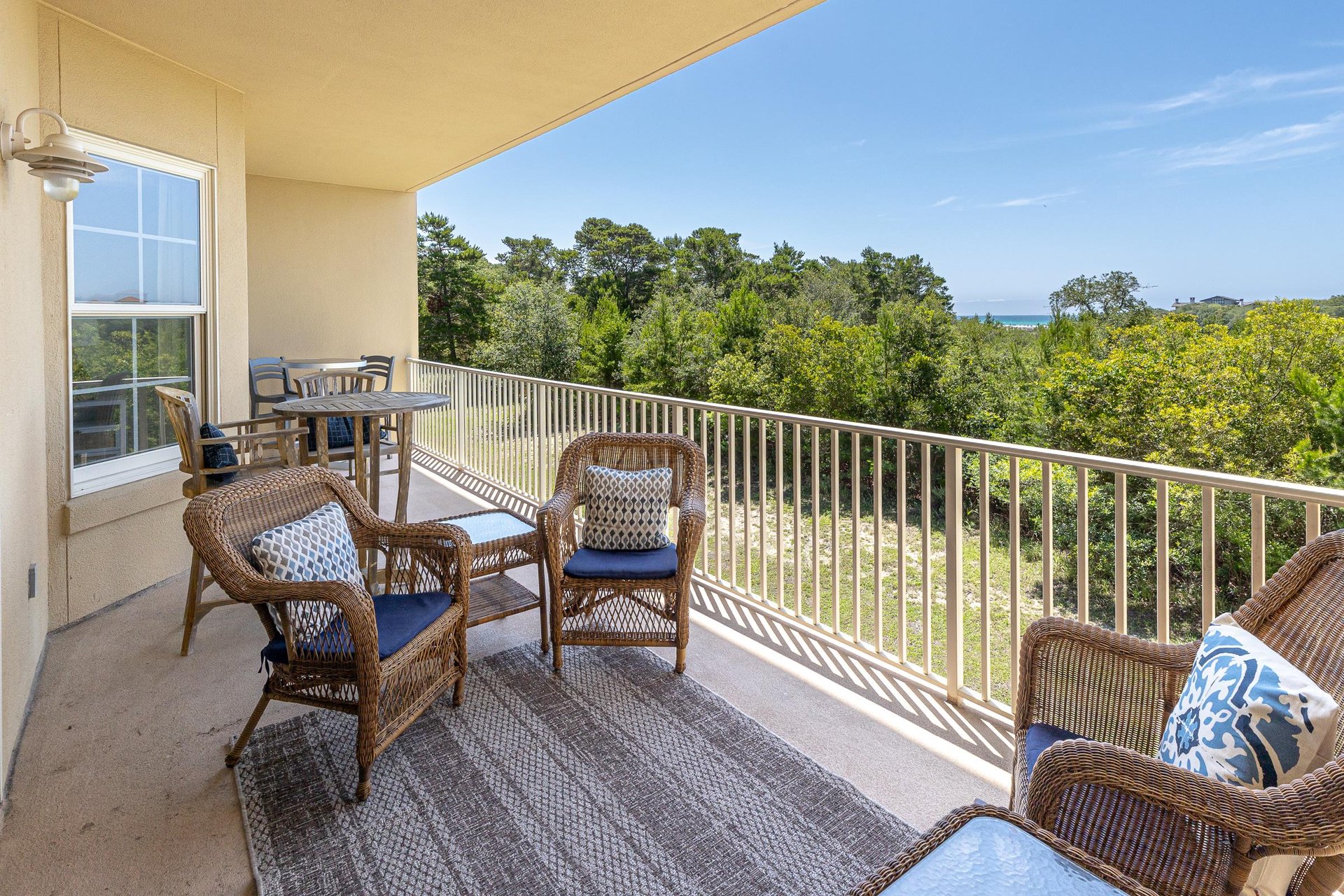 Expansive balcony overlooking lush treetops and distant sea views, featuring wicker seating with blue cushions and a dining table—perfect for morning coffee or sunset relaxation.