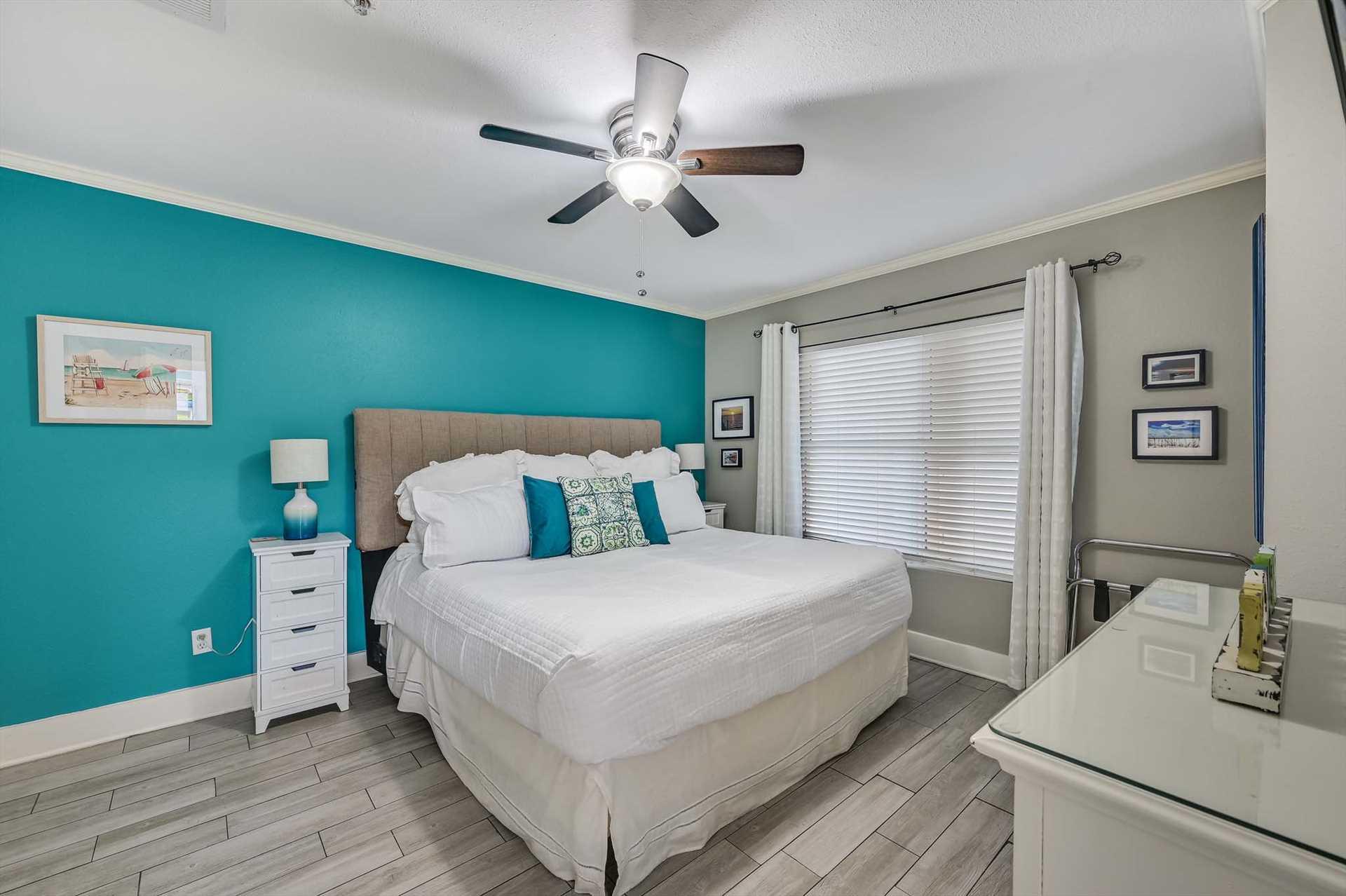 Step into Bedroom 2 providing bright, coastalinspired bedroom with a plush bed and soothing turquoise accent wall. The ceiling fan keeps you cool while natural light floods through the window.