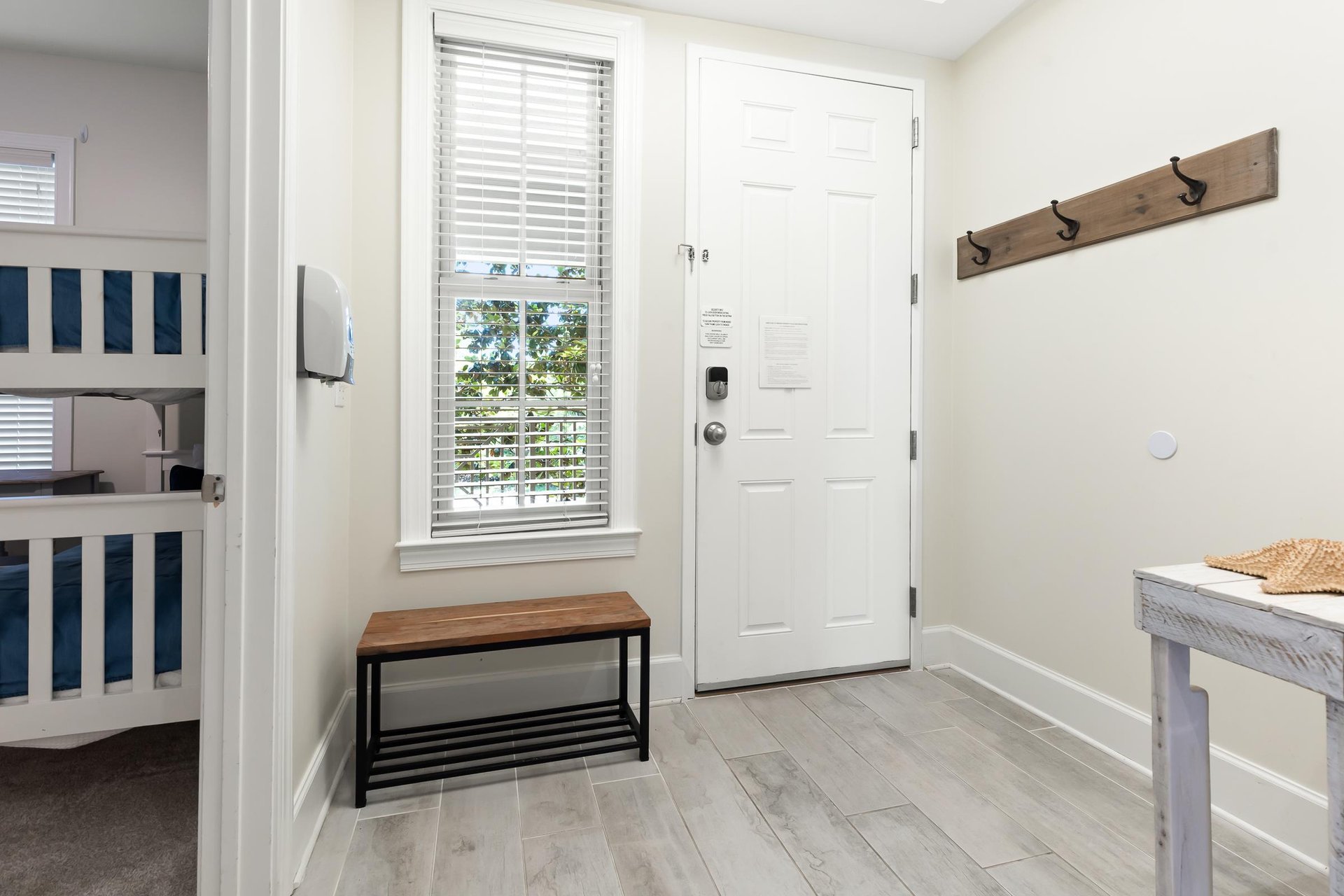 A bright entryway greets you with coastal charm—wooden hooks for beach bags, a bench for sandy feet, and glimpses of a cozy bunk room ready for family fun.