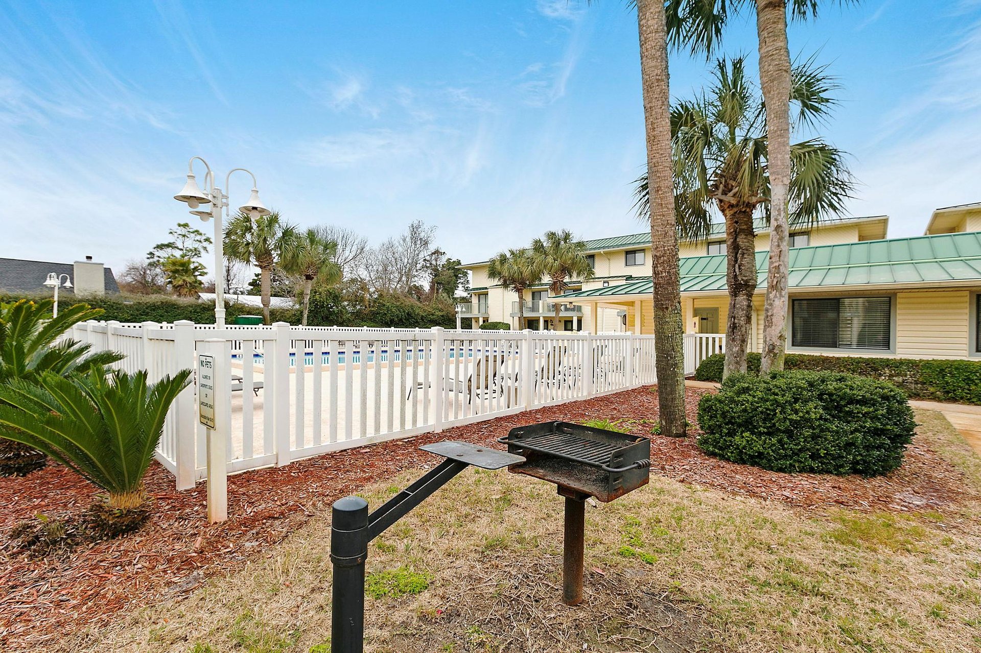 The property features shared outdoor spaces with BBQ facilities, surrounded by tropical palms and coastal landscaping, offering easy access to nearby amenities.