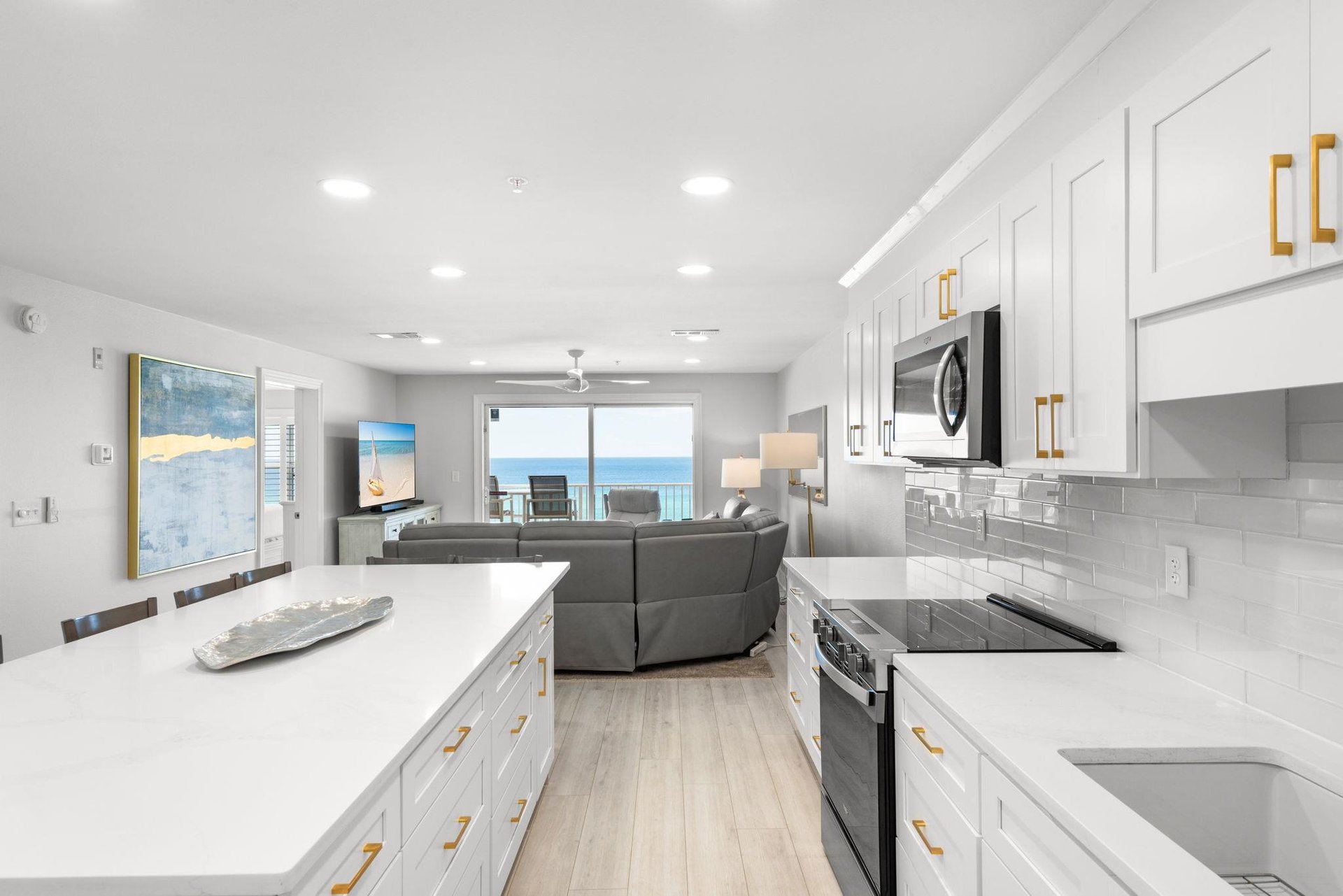 Bright, modern kitchen with sleek white cabinetry and gold hardware opens to stunning ocean views. Sunlight streams through the balcony doors, perfect for coastal living.