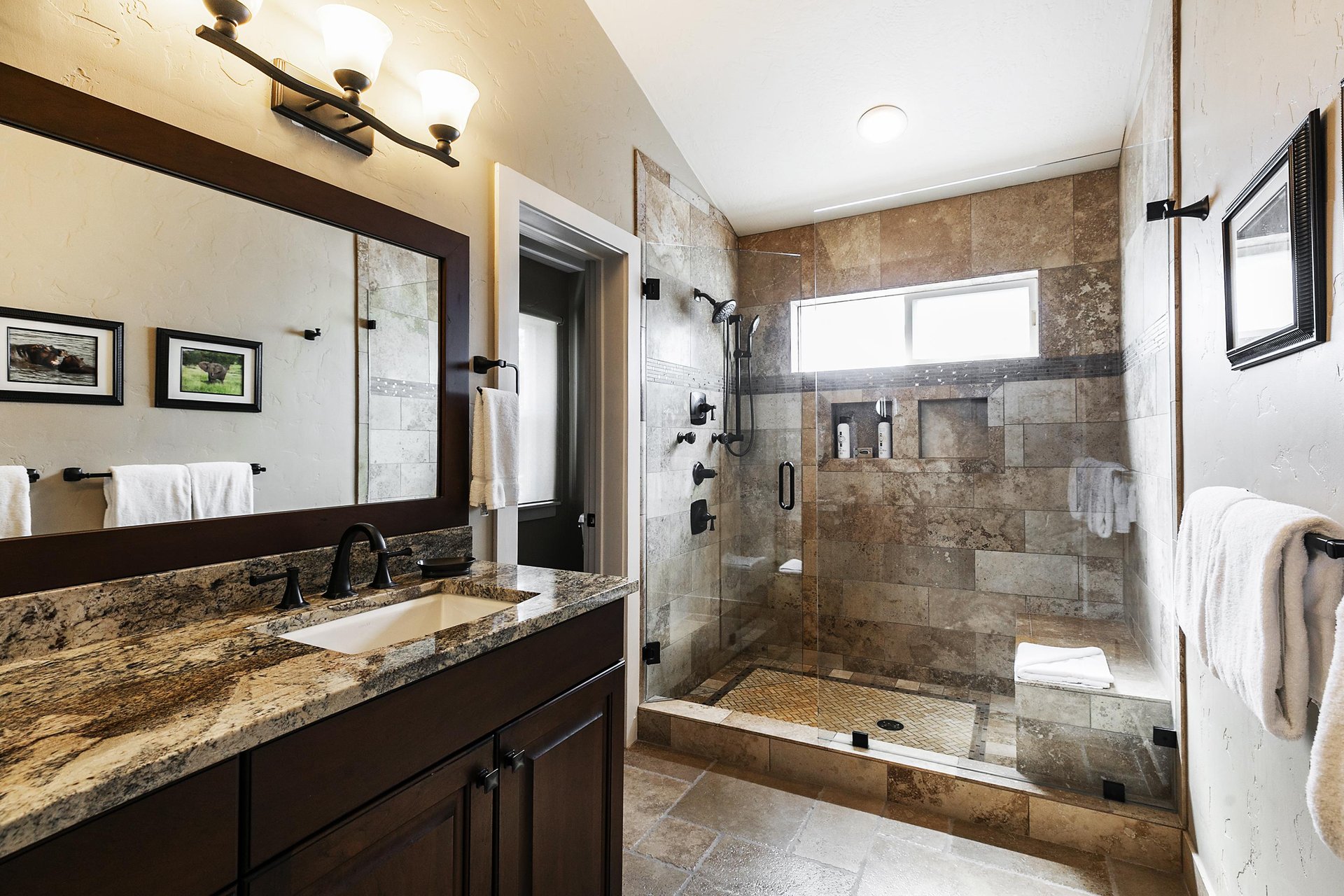 Main bathroom with vanity lighting, granite countertops, wood cabinetry, and tiled shower