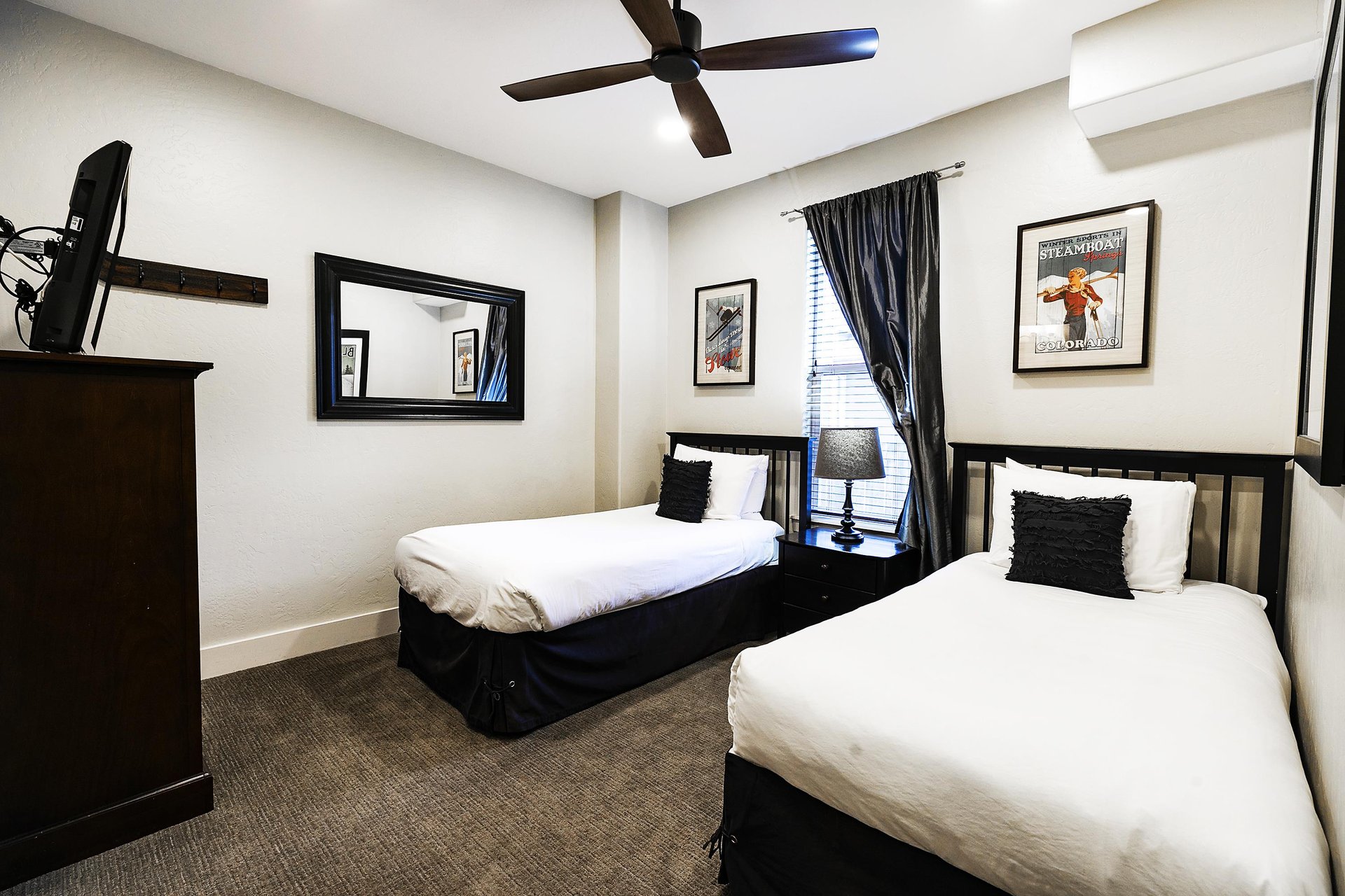 Guest bedroom with two twin beds, white linens, picture window with dark drapes, wooden dresser with TV, and carpeted floor