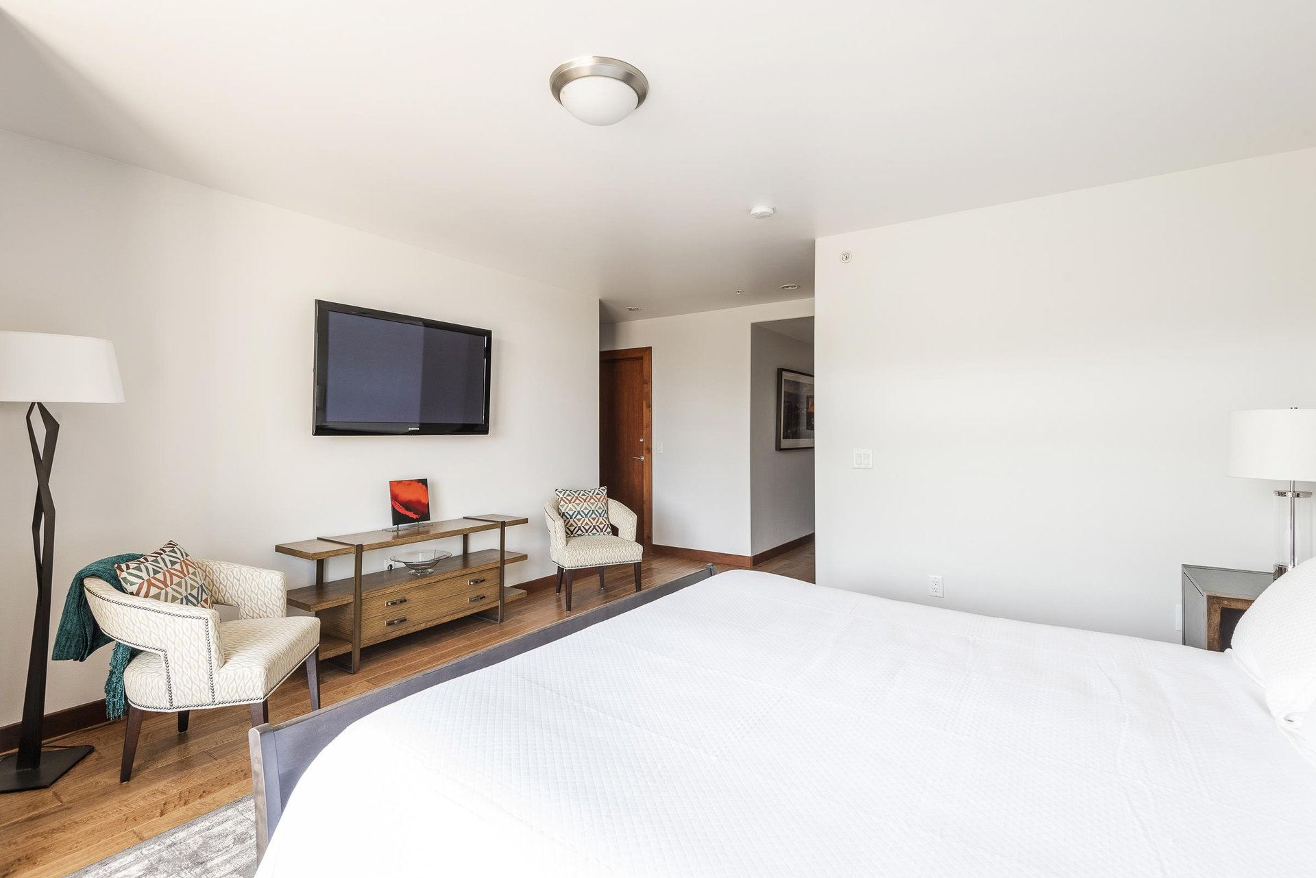 King bed in the main bedroom looking toward the large screen wall mounted TV over a wooden accent table with 2 armchairs