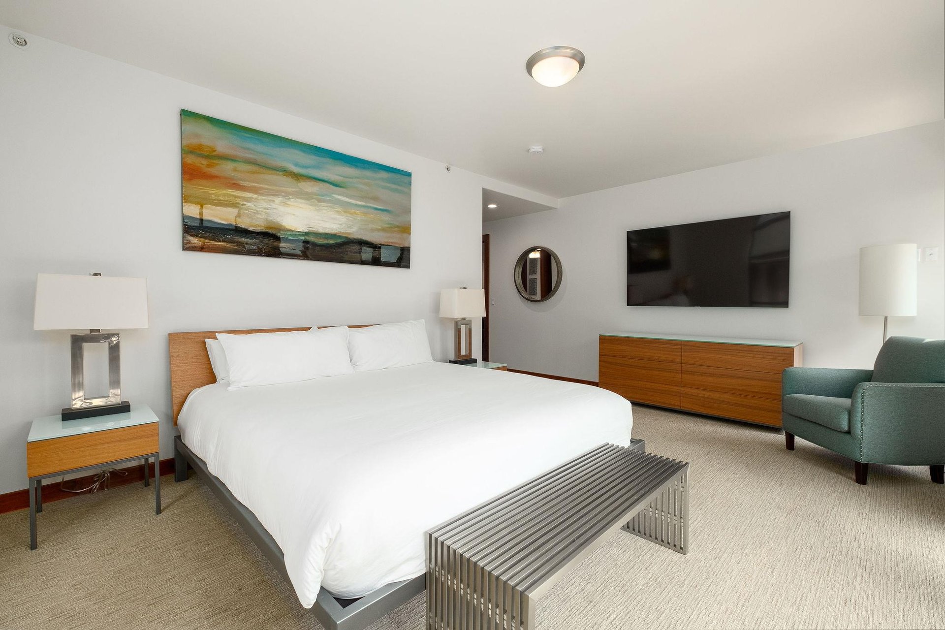 Luxurious platform king bed in the main bedroom draped in white linens and duvet, wooden headboard and accent pieces and large wall mounted flatscreenTV