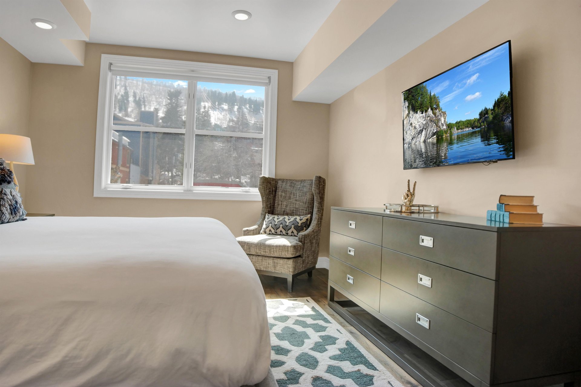 Black chest of drawers, flat screen TV and upholstered sitting chair next to the large window with mountain views.