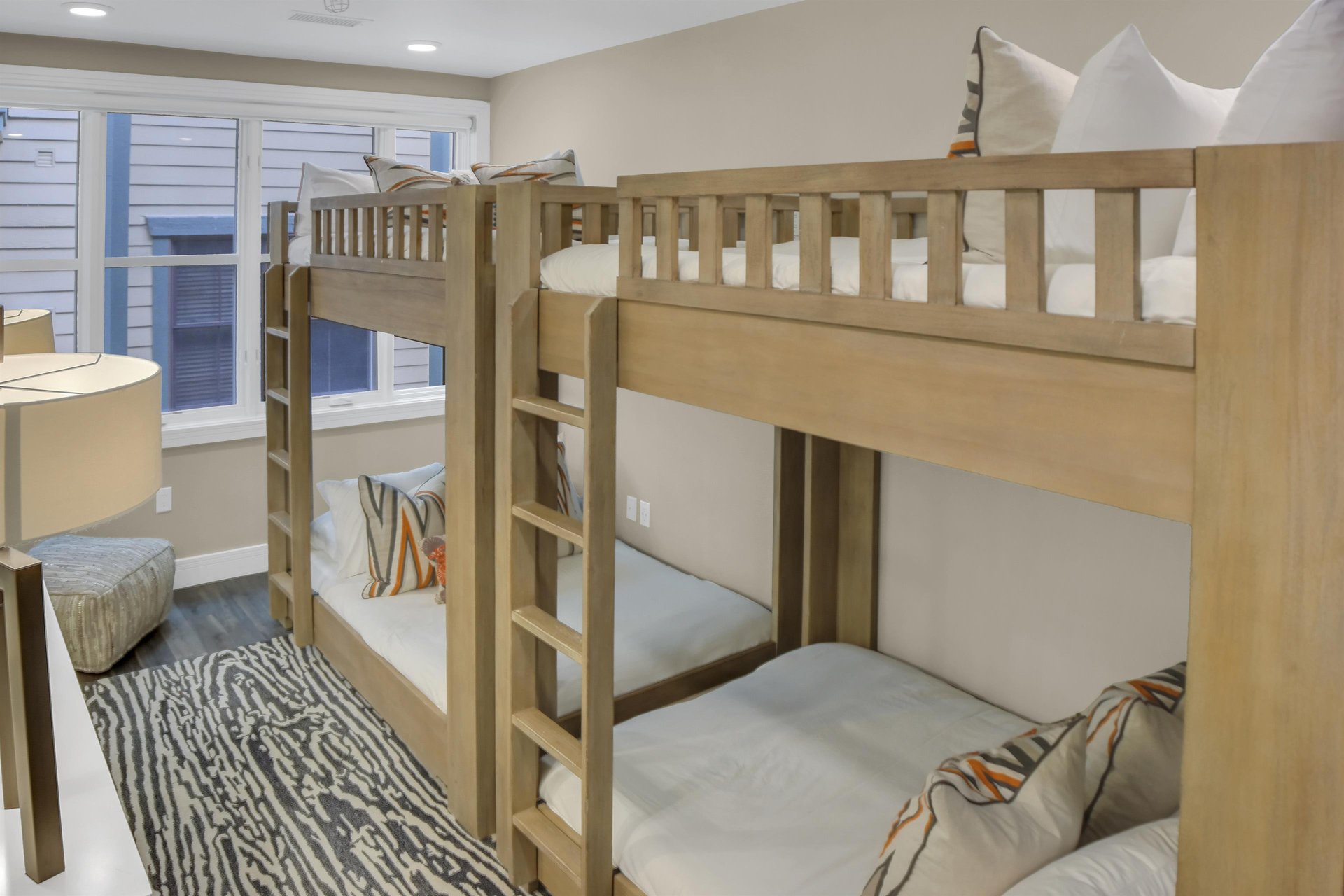 Two light colored wood bunkbeds, pushed end to end with orange and grey zig zag throw pillows and large window.