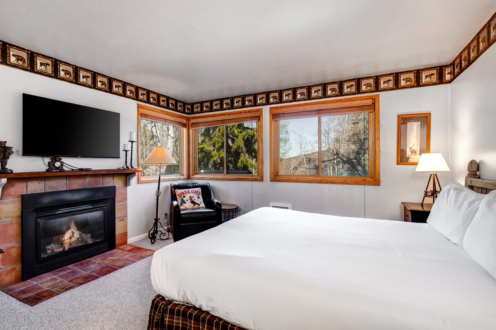 Snow Flowers main suite has a king size bed with white linens, fireplace and TV, blue chair under 3 windows and floor lamp.