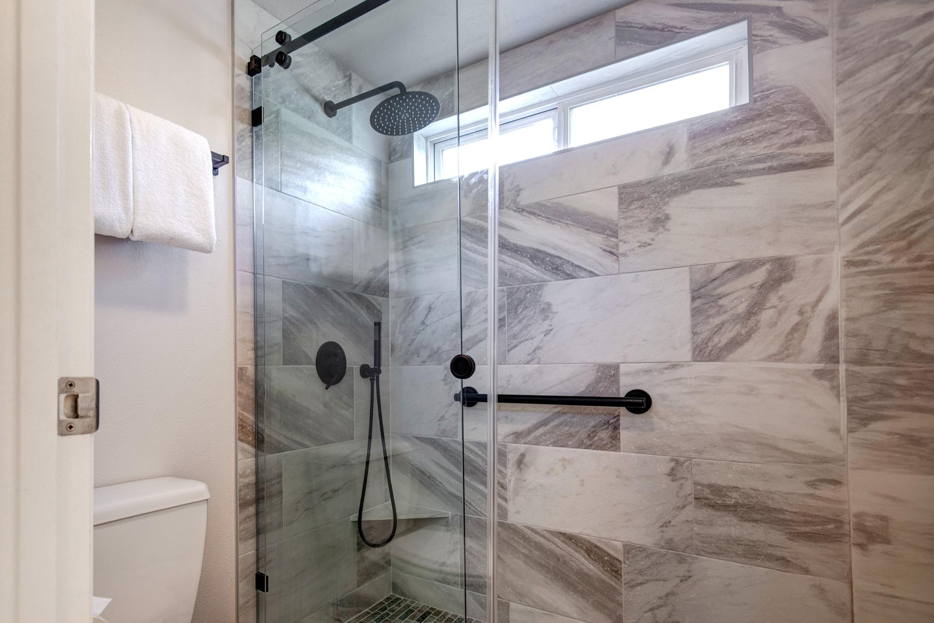 The walkin shower has grey and white patterned wall tile, black hardware, white toilet, and a white folded hanging towel.