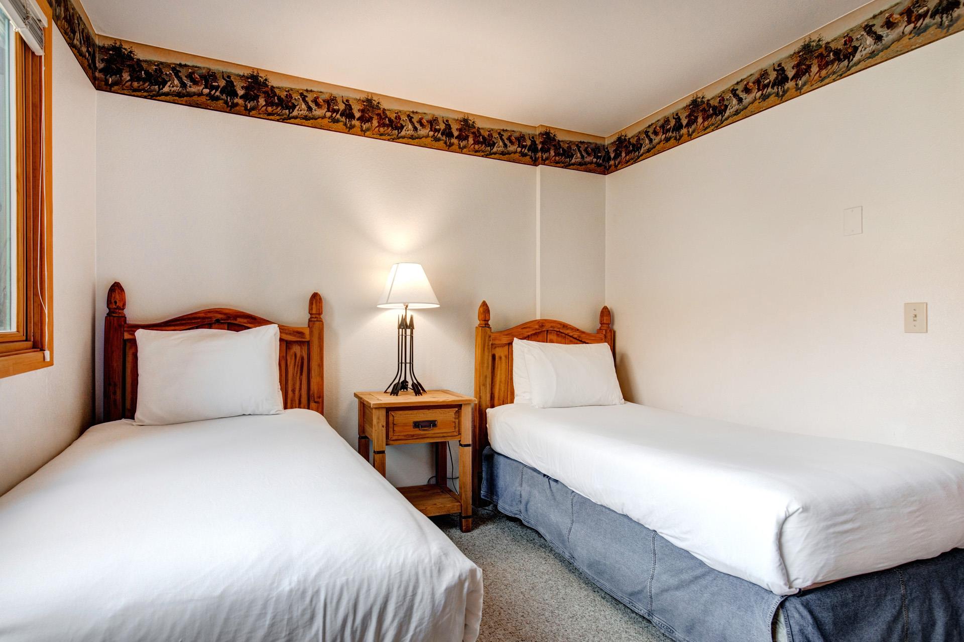 Two twin beds with medium toned wood headboards, blue bed skirt, and a nightstand and light in between them.