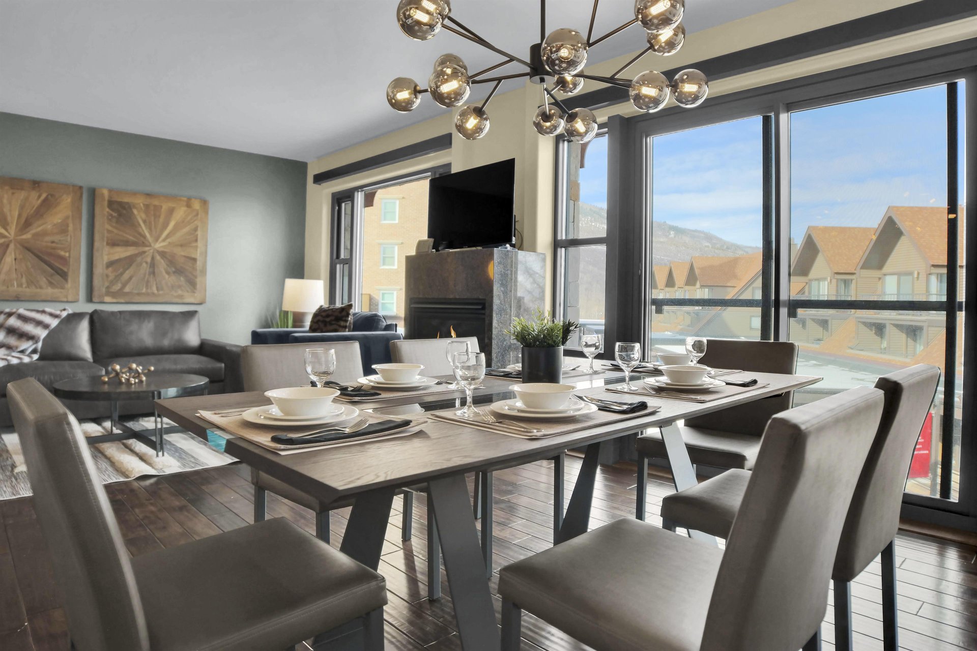 A modern dining table with 6 chairs and place settings, large windows and sliding doors leading to a patio, and chandelier.