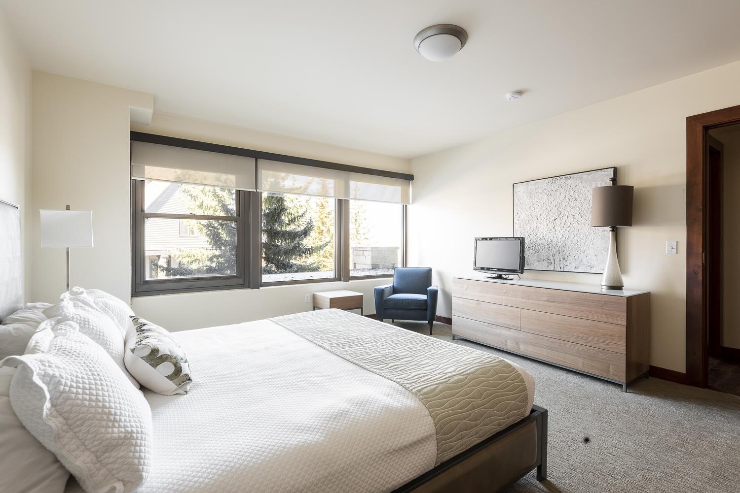 Luxurious king bed in the main suite, large picture windows overlooking the plaza, wooden dresser with TV and window side lounge chair