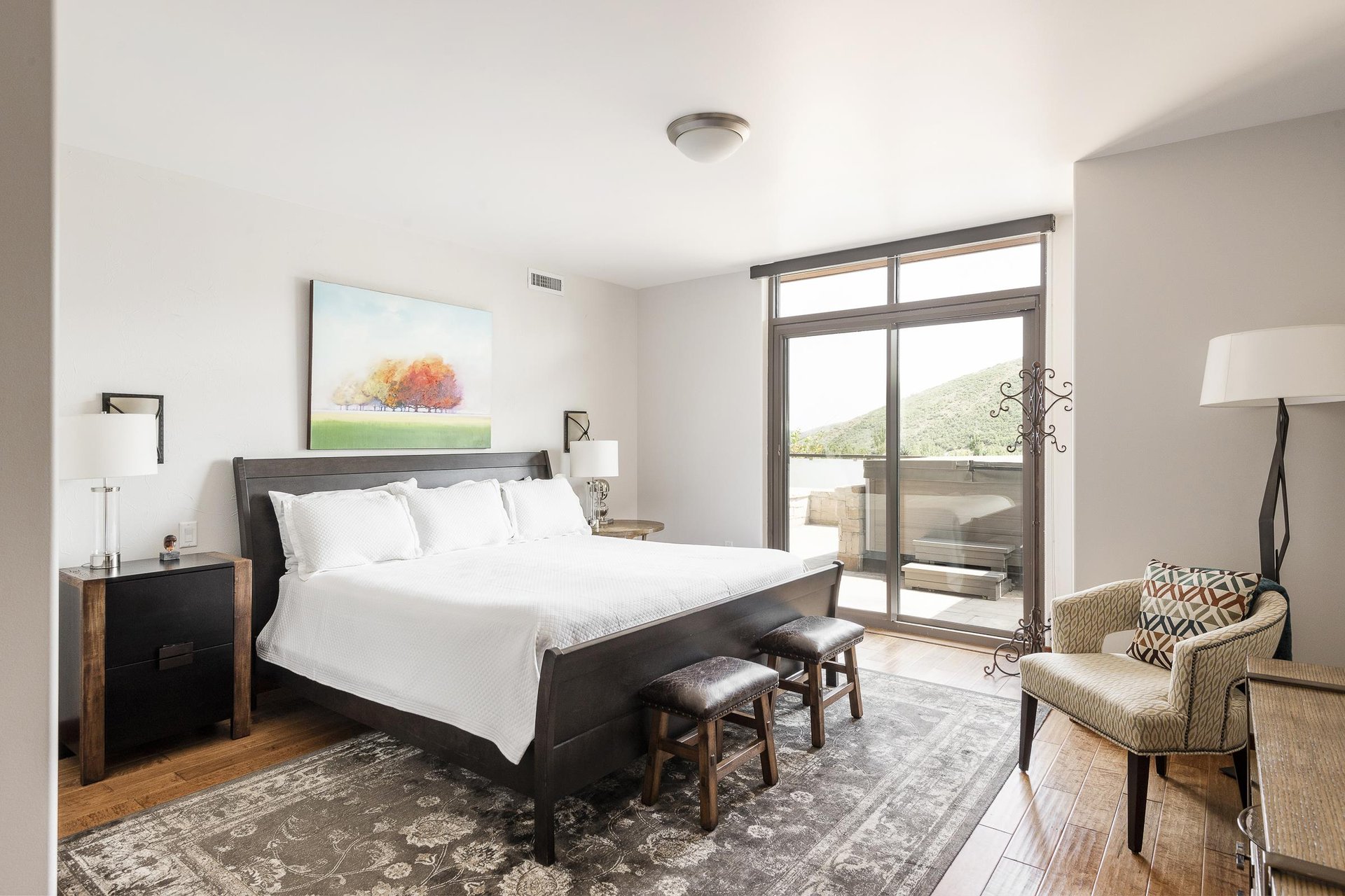 Main suite king bed, white linens and duvet, wooden headboard, hardwood floors with large Persian area rug, and sliding patio doors to access private deck