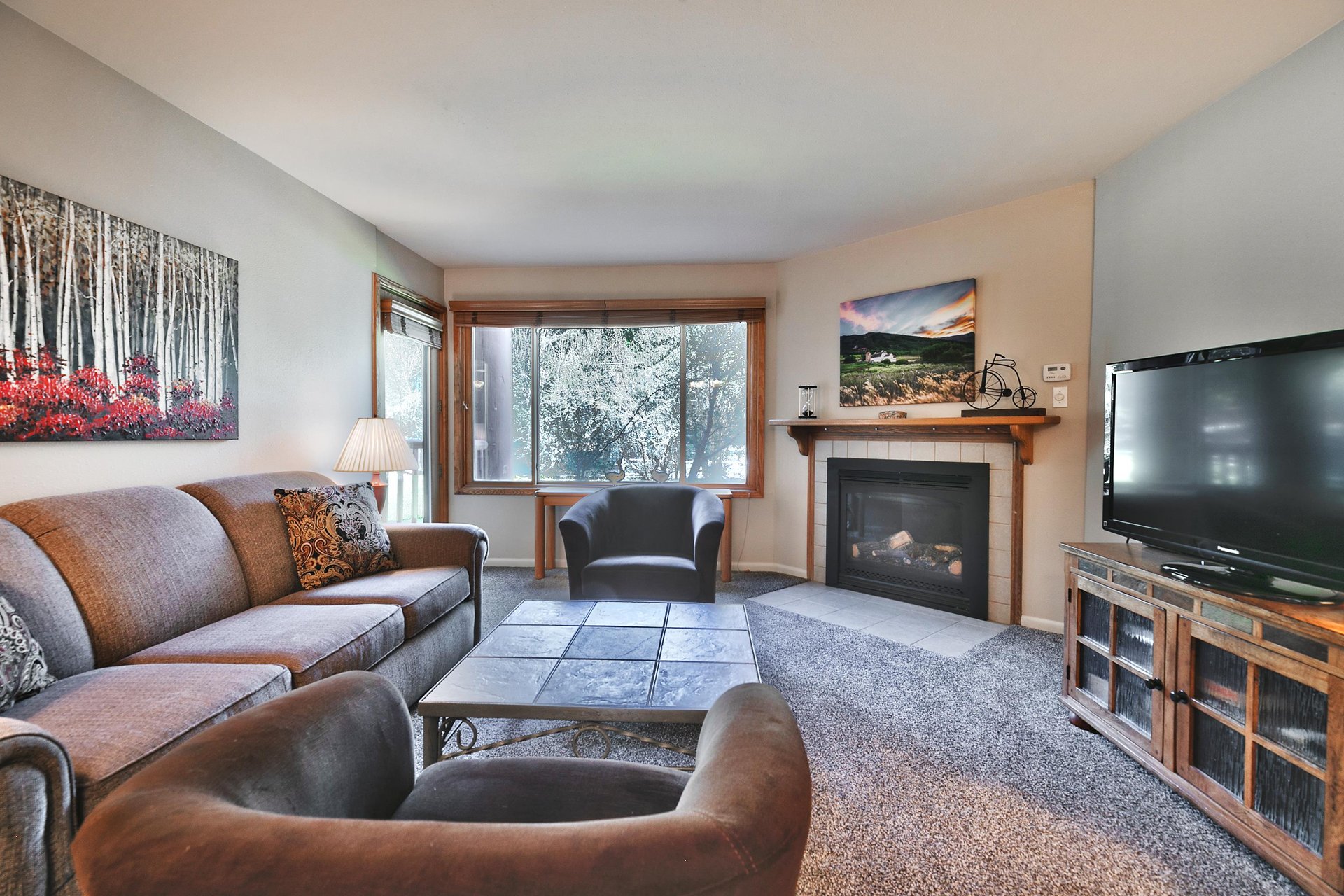 Living room includes a gas fireplace with white tile surround and views of the tennis courts from the picture window