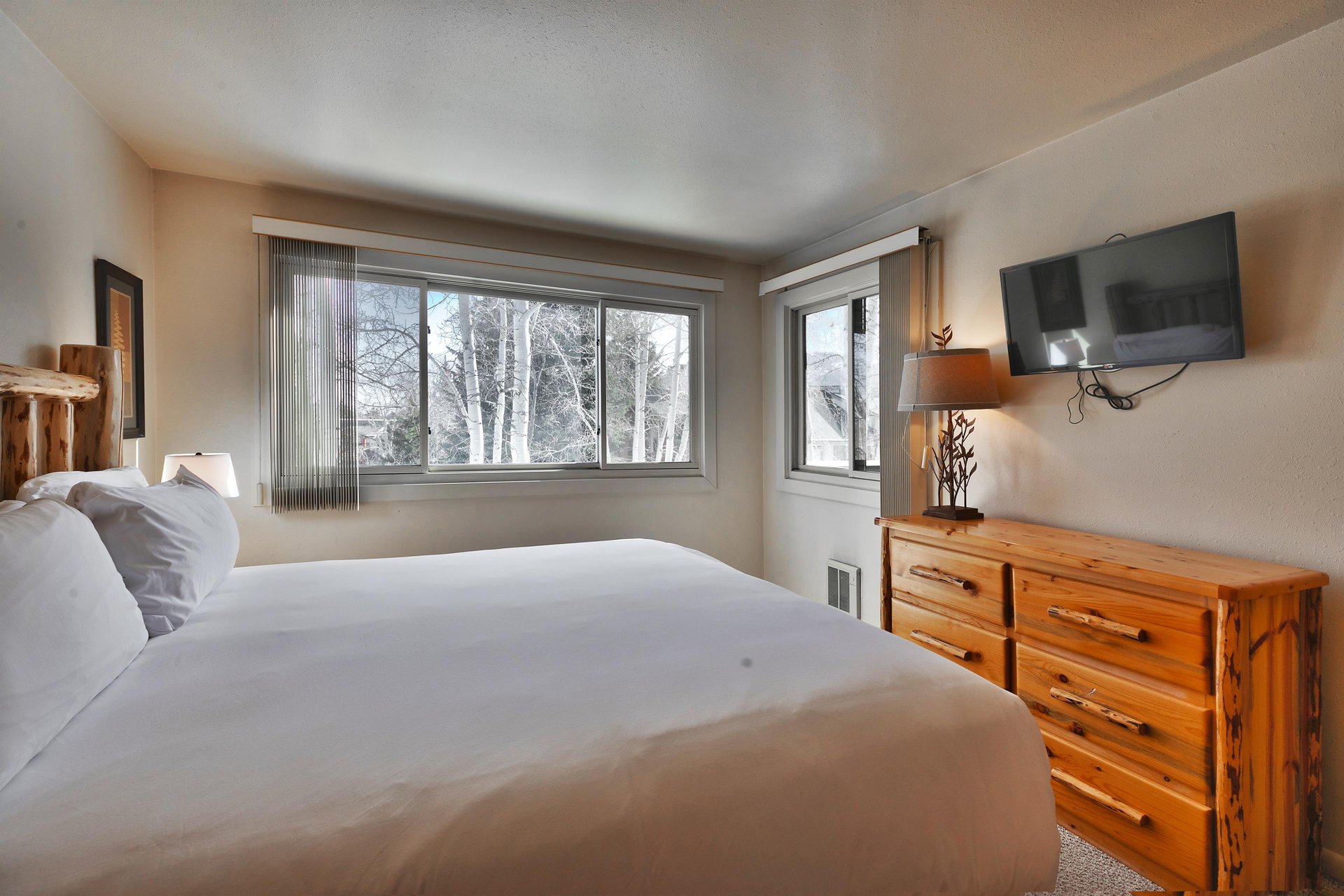 The main bedroom has an abundance of light from the windows, a wall mounted flat screen TV, beautiful lamps and a log dresser