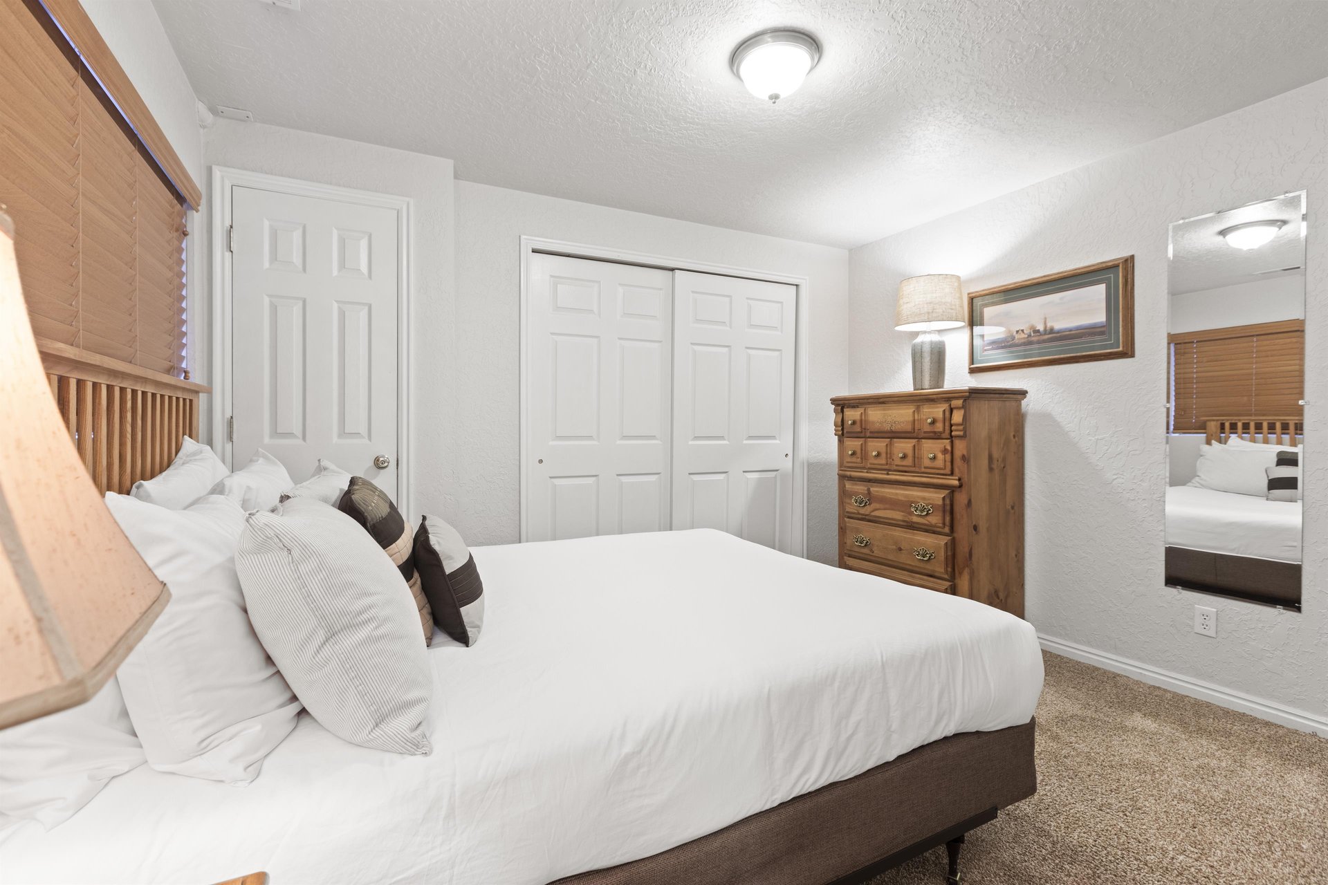 A bedroom with a queen size bed, wooden dresser and white walls and doors.