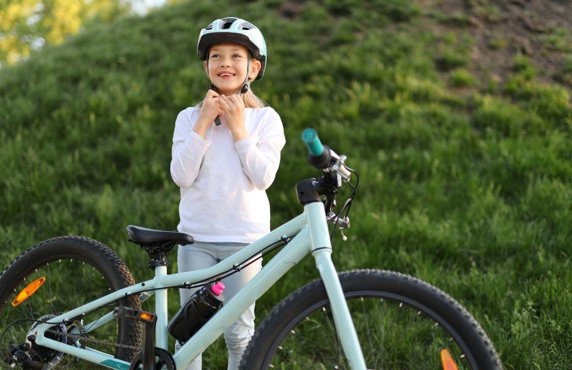 Young girl fastens her bike helmet while standing next to her powder blue mountain bike.