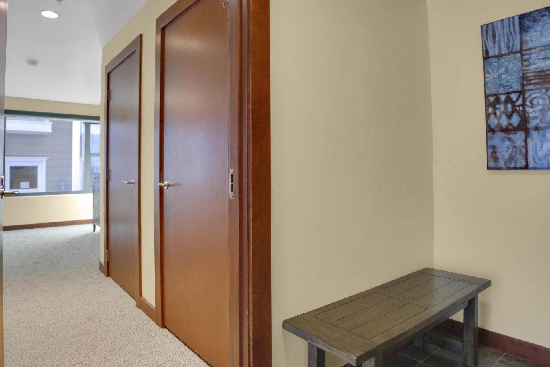 The entryway with a bench, and a hall with two doors, cream colored carpet and creamy yellow colored walls.