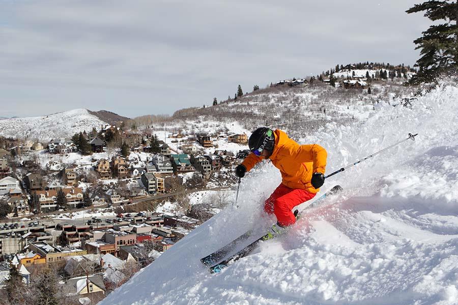 A skier dressed in orange and red at Park City Mountain Resort on a steep snowy slope with the town behind him.