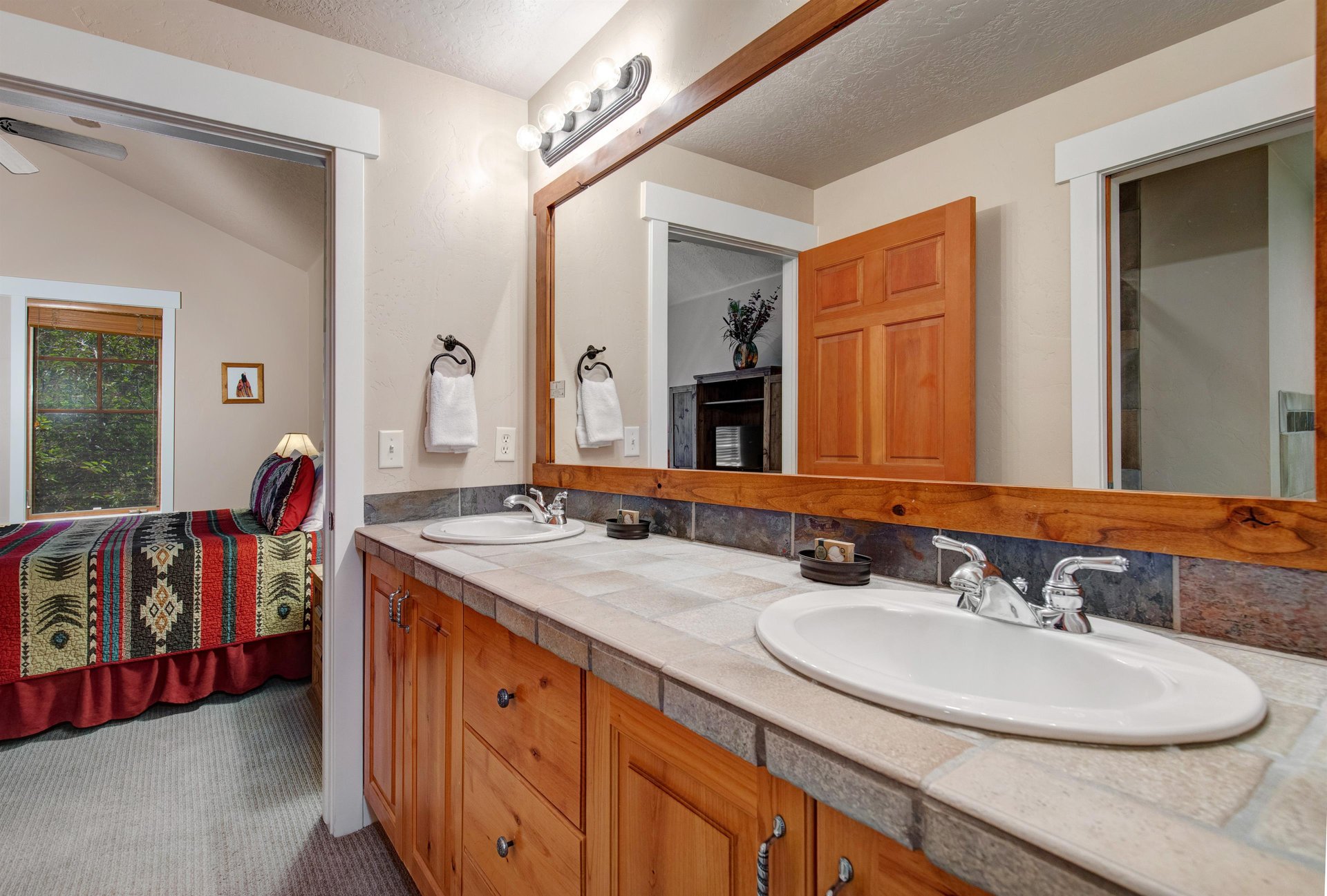 Spacious dualsink bathroom with warm wood cabinetry and tile countertops. Fresh towels await your arrival, and the adjacent bedroom provides a cozy mountain retreat.