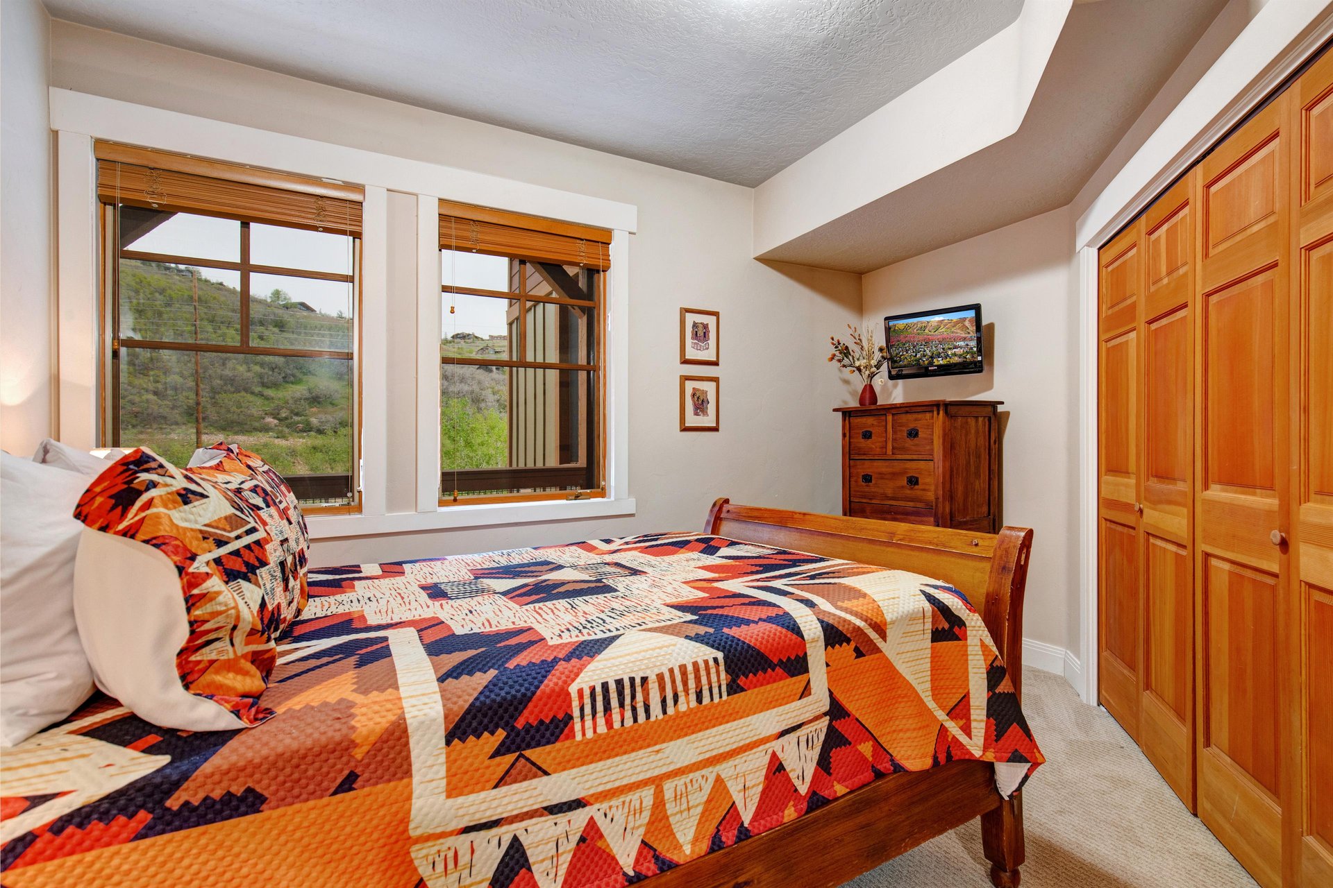 Settle into your cozy bedroom with rich wood tones, a vibrant quilt, and windows framing natural hillside views. Your dresser holds a flatscreen TV, and your wardrobe offers ample closet space for un