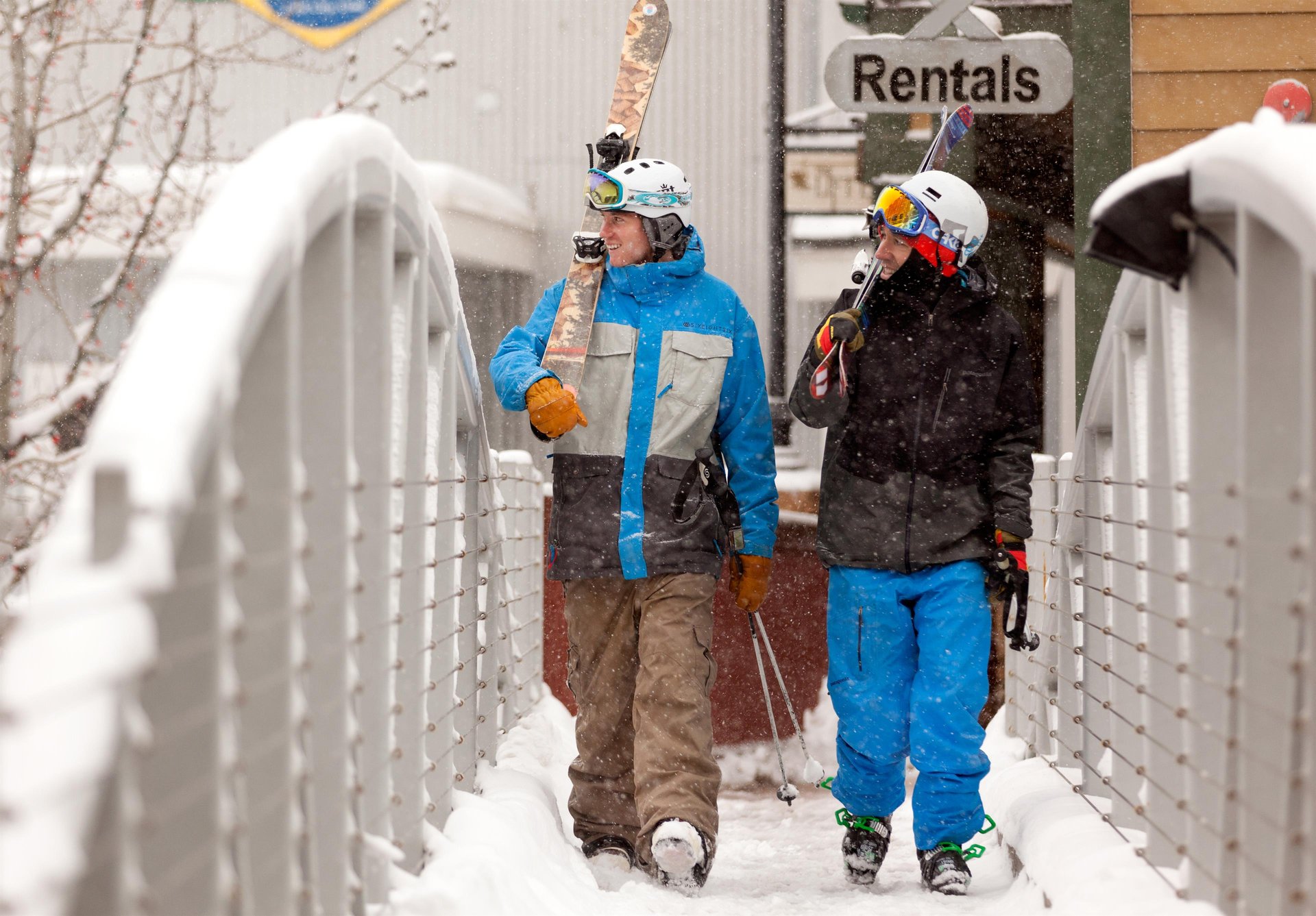 Two skiers equipped with rental gear pause on a snowy bridge near a rental shop, ready for a winter day on the slopes.