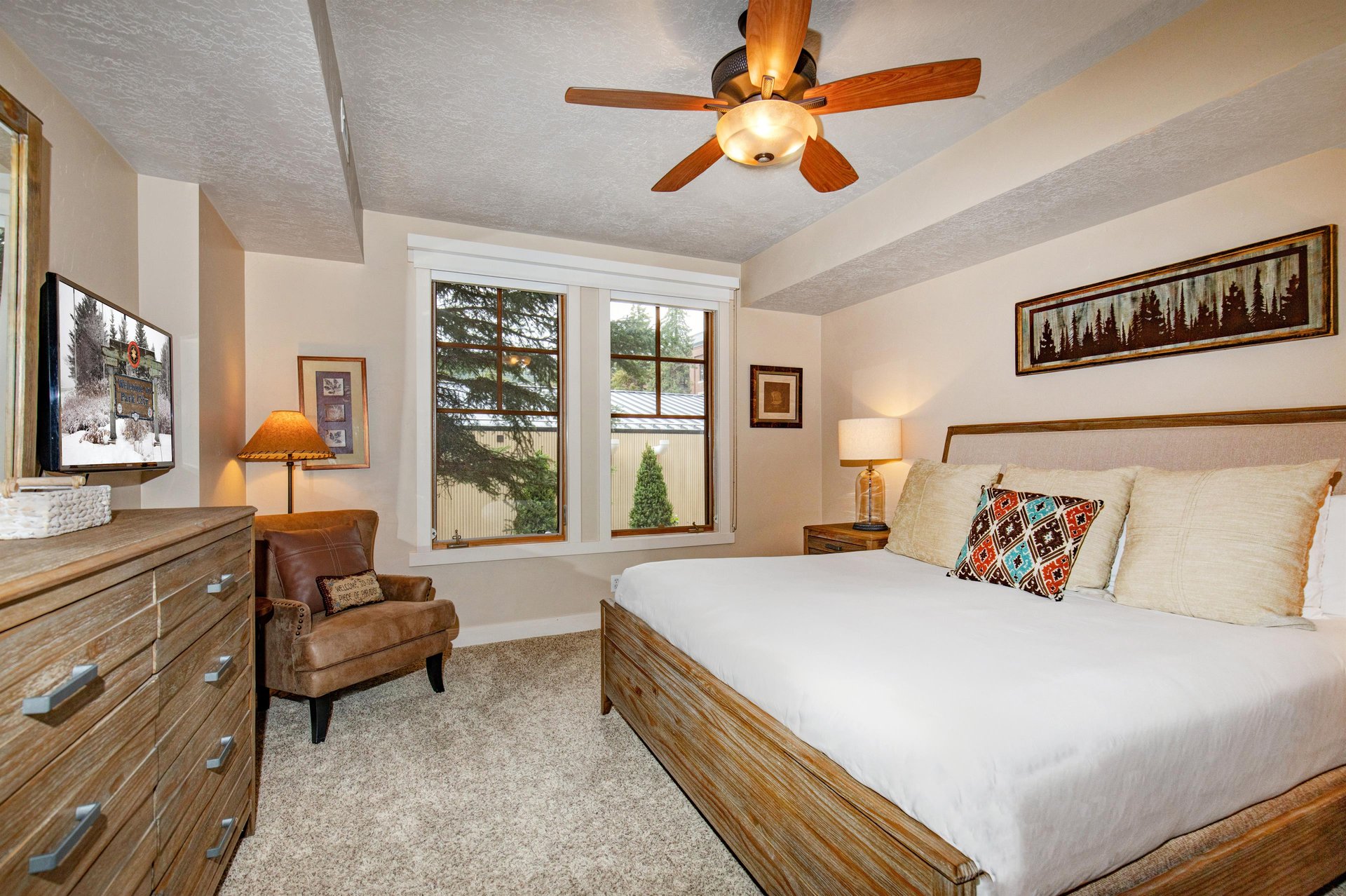 Your peaceful retreat features a plush queen bed, cozy armchair, and ceiling fan for comfort. Natural light filters through the window while warm wood tones create a serene mountain escape.