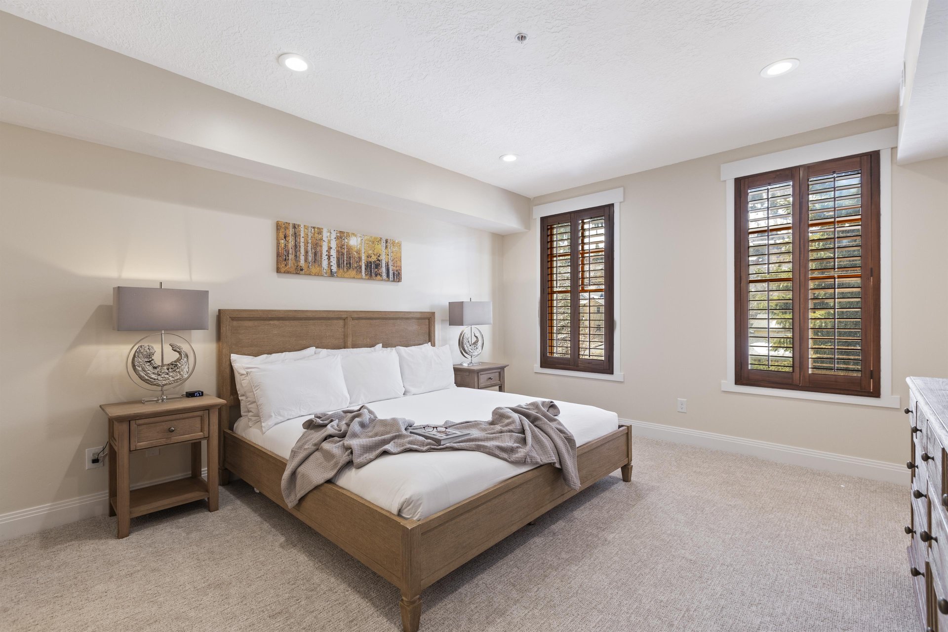 Main bedroom with king bed, wood nightstands, reading lamps, large windows and warm natural light.