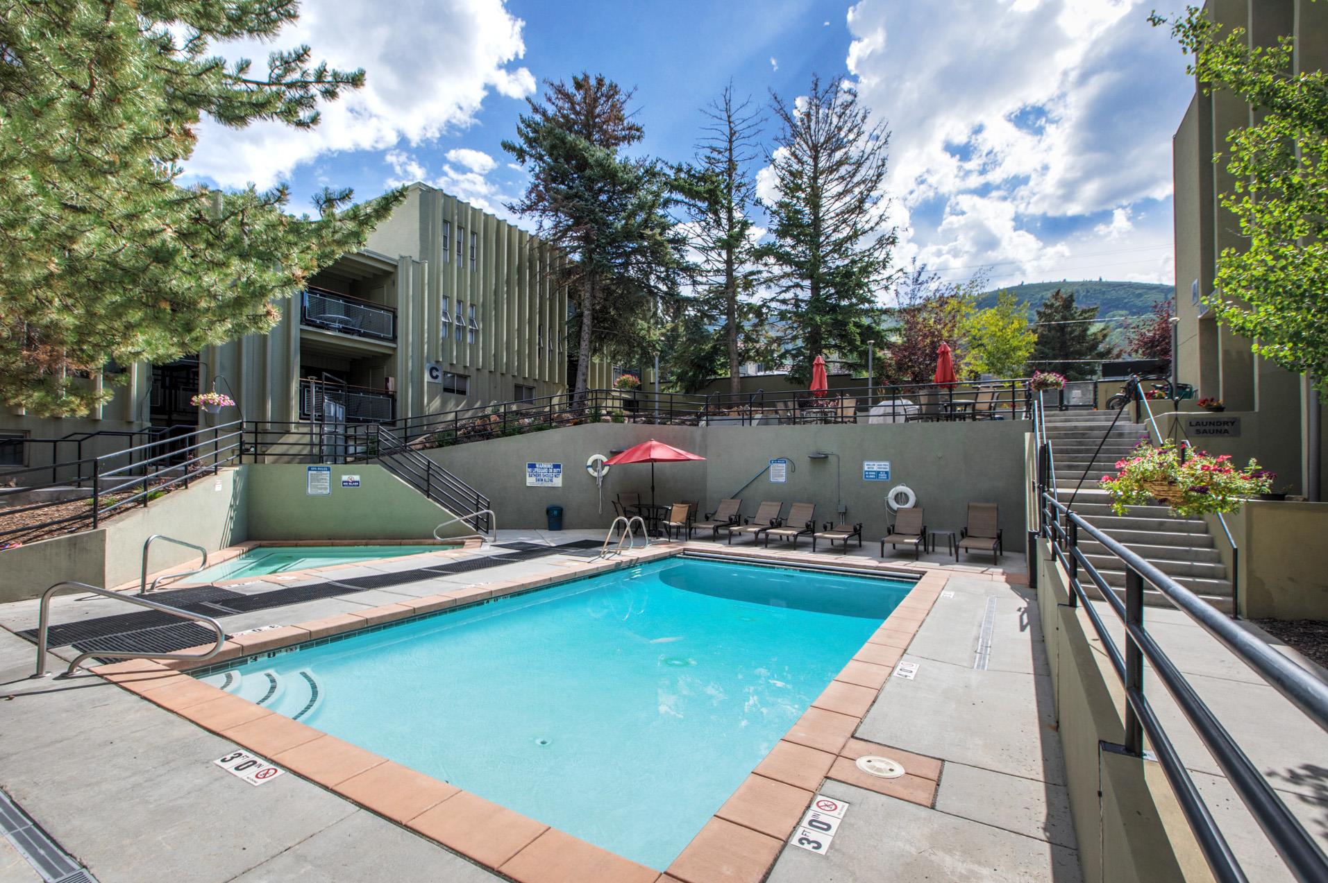 Outdoor community pool surrounded by lounge seating, mature trees and mountain scenery.