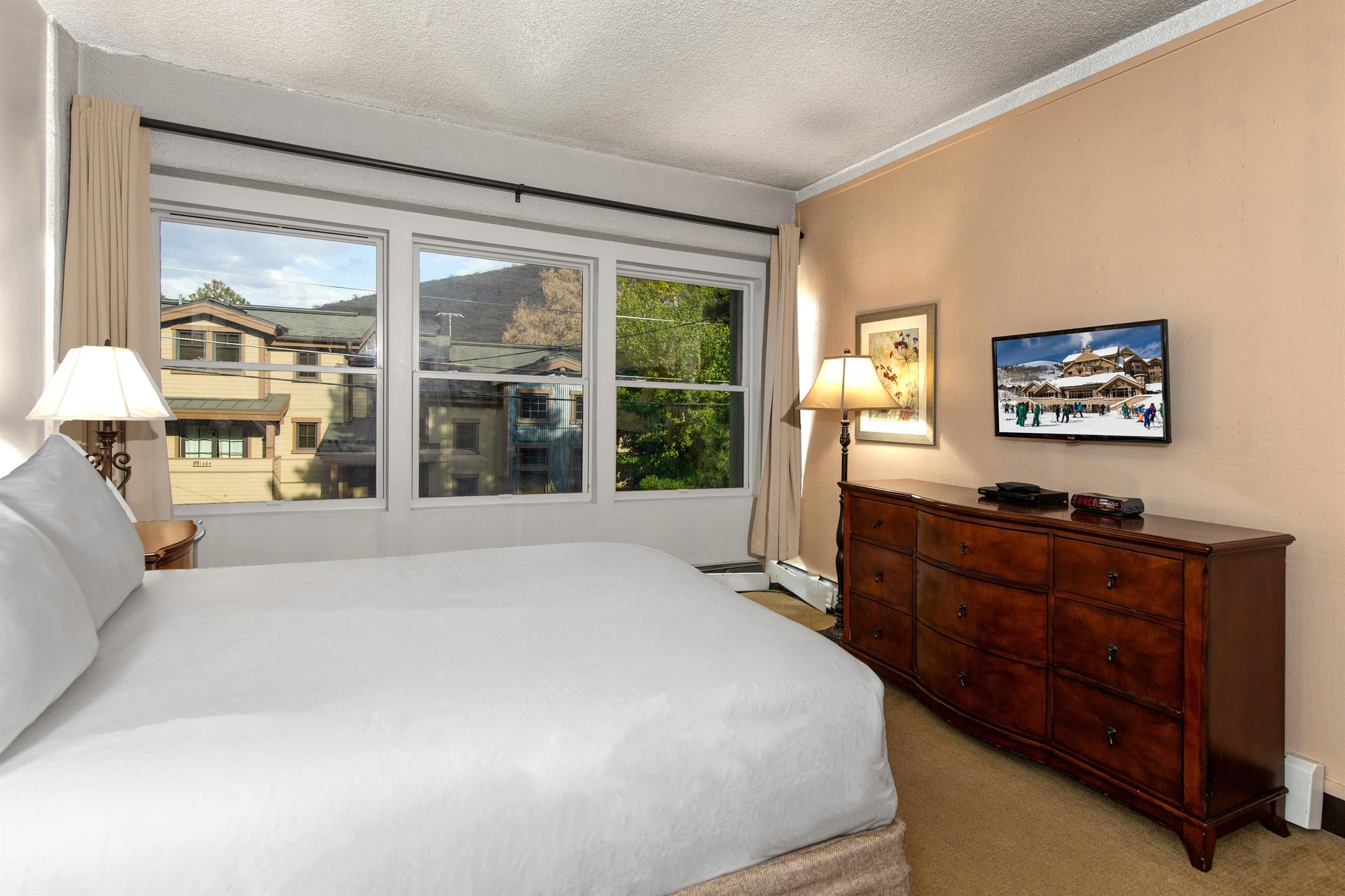Bright bedroom with queen bed, wood dresser, TV and large windows overlooking the surrounding mountain area.