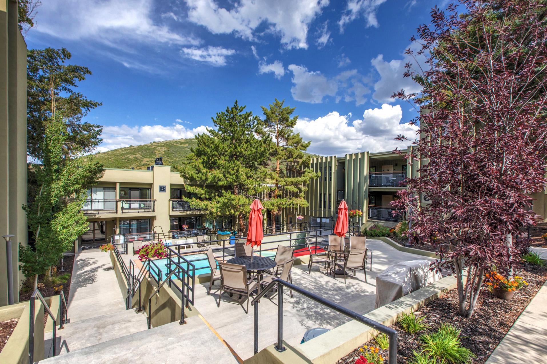 Community outdoor pool and patio area with lounge seating, tables, umbrellas and surrounding trees.