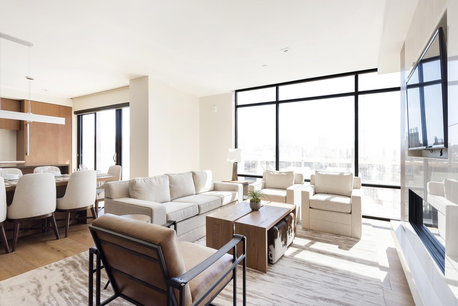 Stunningly bright living room with striking window walls, hardwood floors and large area rug, white couch, two white arm chairs, and leather and black metal accent chair.