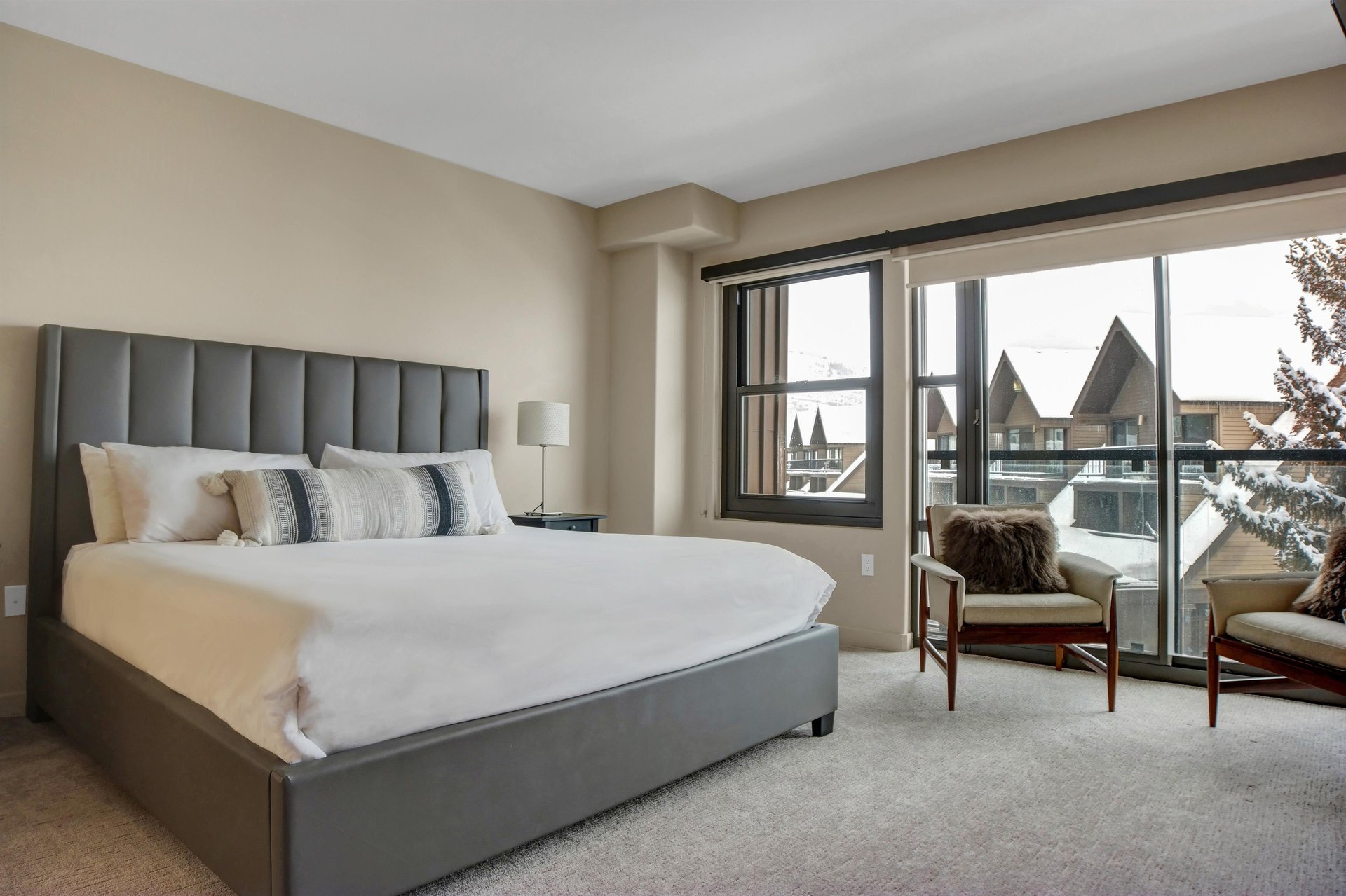 Bright bedroom with king bed, upholstered headboard, sitting chair and large windows with mountain views.