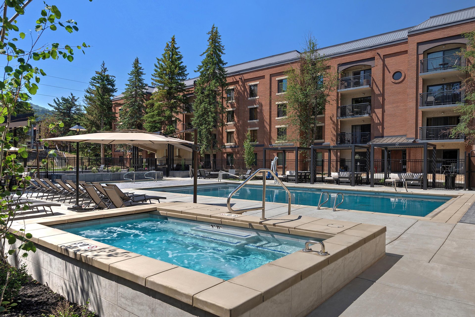 Outdoor community pool and hot tub with lounge chairs and surrounding trees.