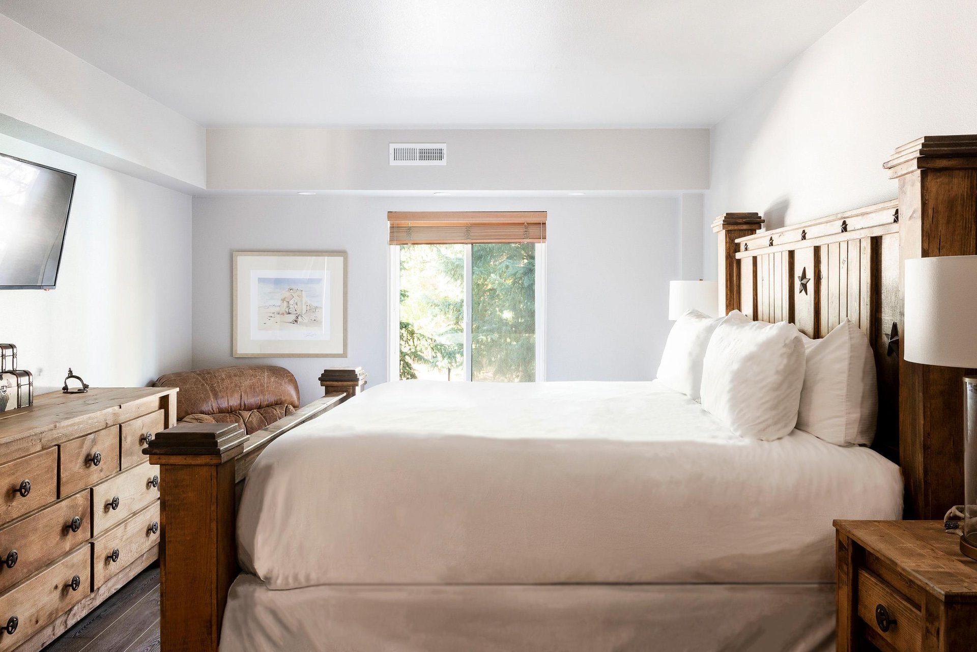 Bright bedroom with white linens, rustic wood bed and large window overlooking the trees.
