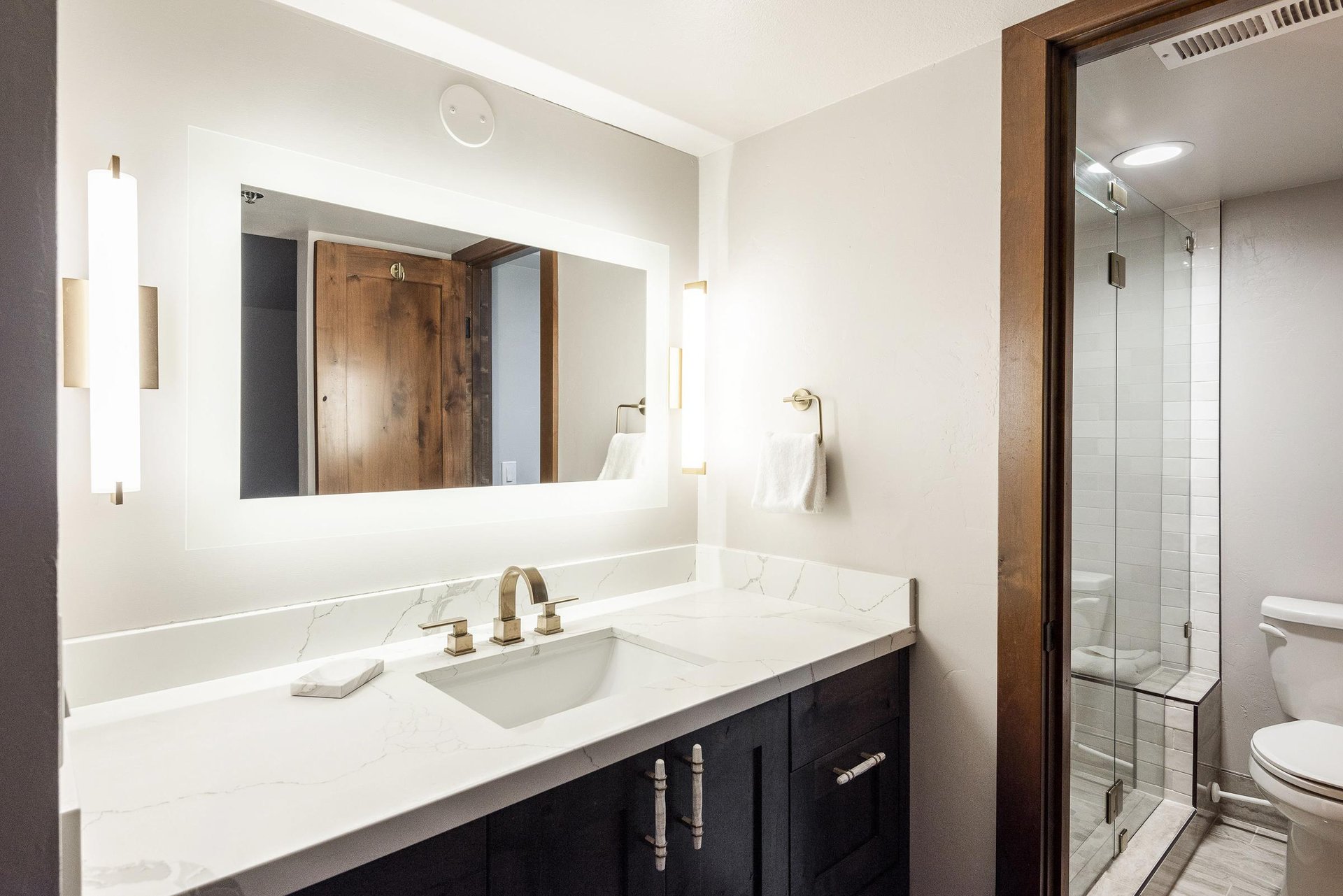 Ensuite bathroom with dark wood vanity, white countertop, large illuminated mirror and glass shower.