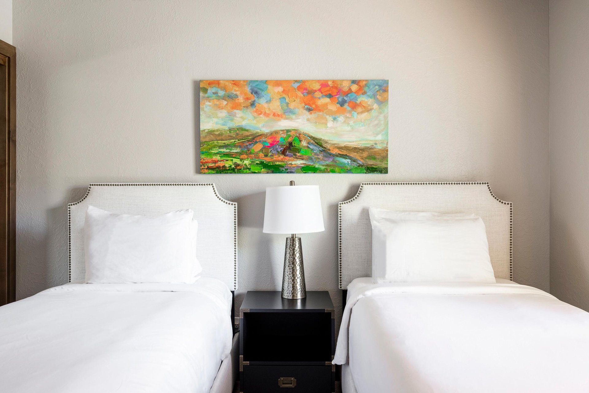 Two twin beds with crisp white linens, shared nightstand, bedside lamp and colorful mountain artwork.
