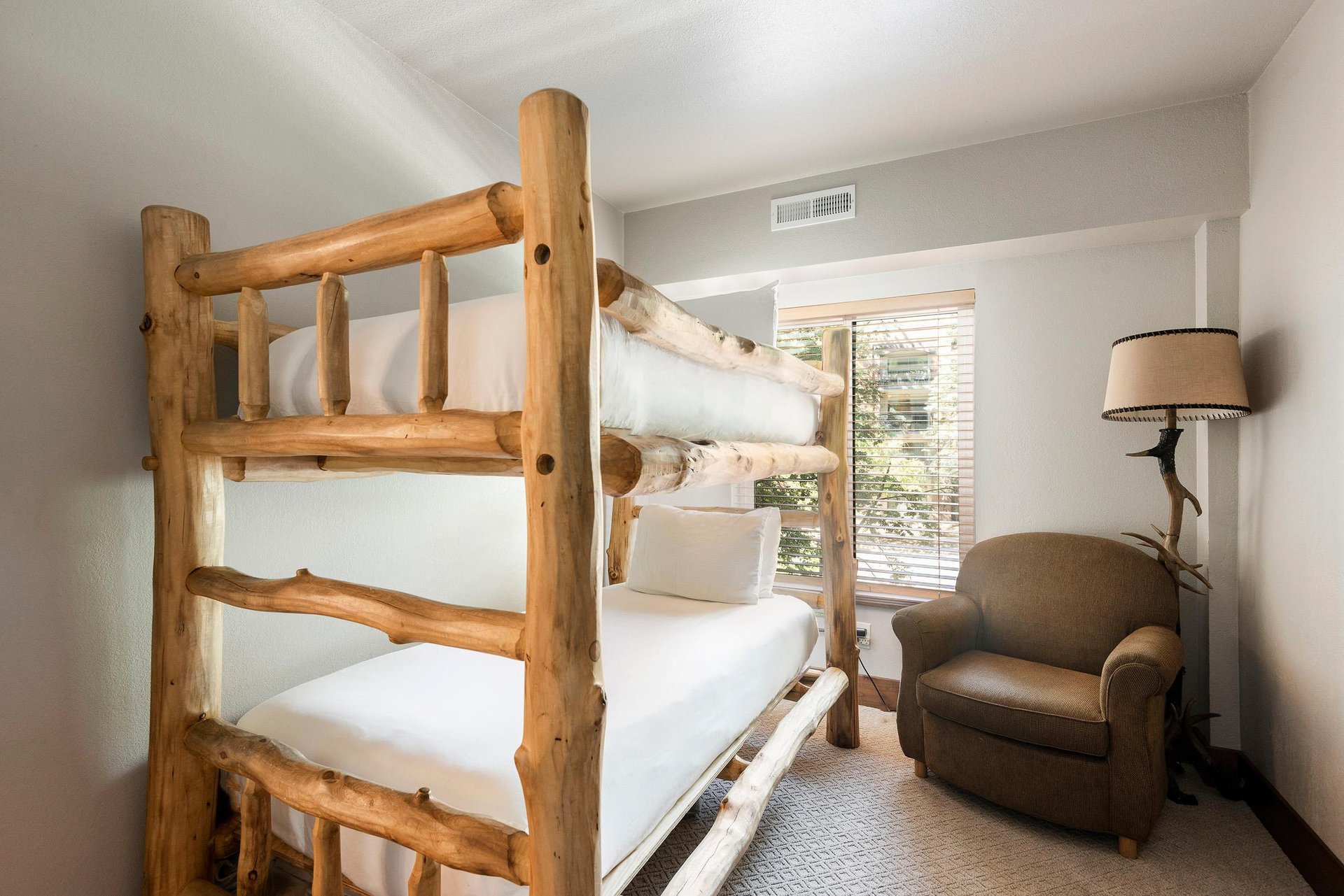 Guest bedroom with rustic log bunk beds, white linens, comfortable armchair and window with tree views.