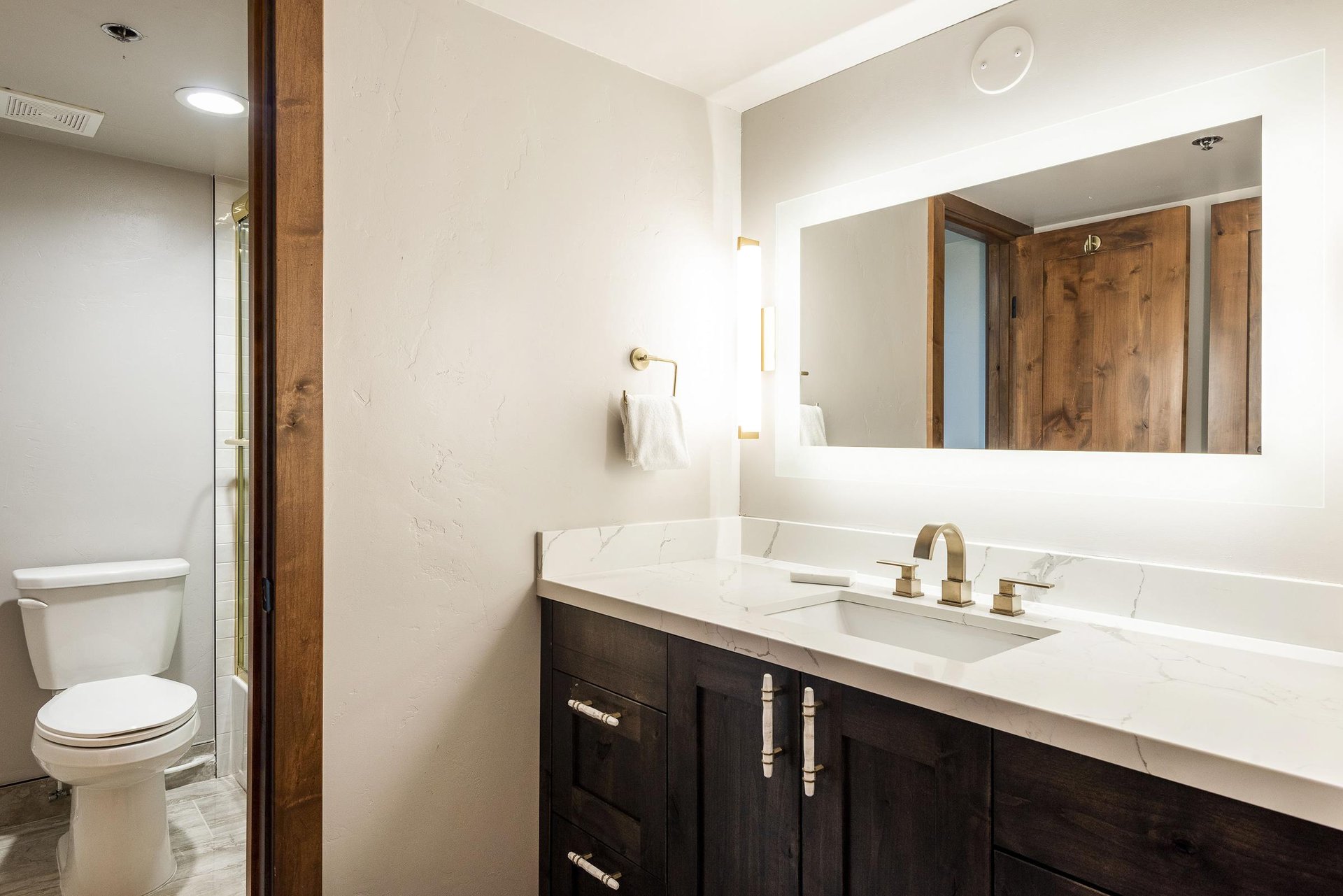Full bathroom with dark wood vanity, white countertop, large illuminated mirror and modern fixtures.