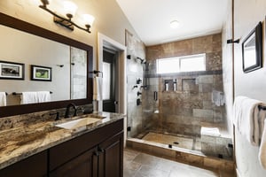 Main bathroom with vanity lighting, granite countertops, wood cabinetry, and tiled shower