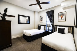 Guest bedroom with two twin beds, white linens, picture window with dark drapes, wooden dresser with TV, and carpeted floor
