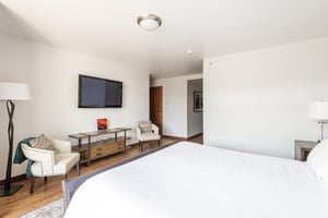 King bed in the main bedroom looking toward the large screen wall mounted TV over a wooden accent table with 2 armchairs