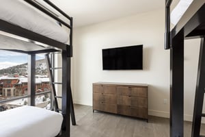Double bunk room  with wooden drawers and wall mounted TV, window wall overlooking the plaza