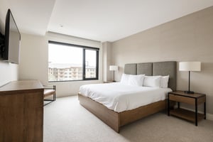 Lush king bed in the main bedroom, wooden platform bedframe with suede headboard, white linens and duvet and large picture window overlooking the plaza