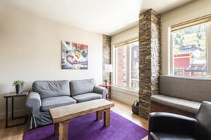 Grey sleeper sofa in the living room with solid wood coffee table, slatestone columns separate the two large windows with a cozy window seat to take in the views of the hillside and Historic Main Stre