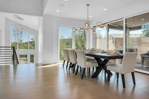 Vacation rental dining room with white chairs, large wooden table, artistic lighting, and surrounded by windows.